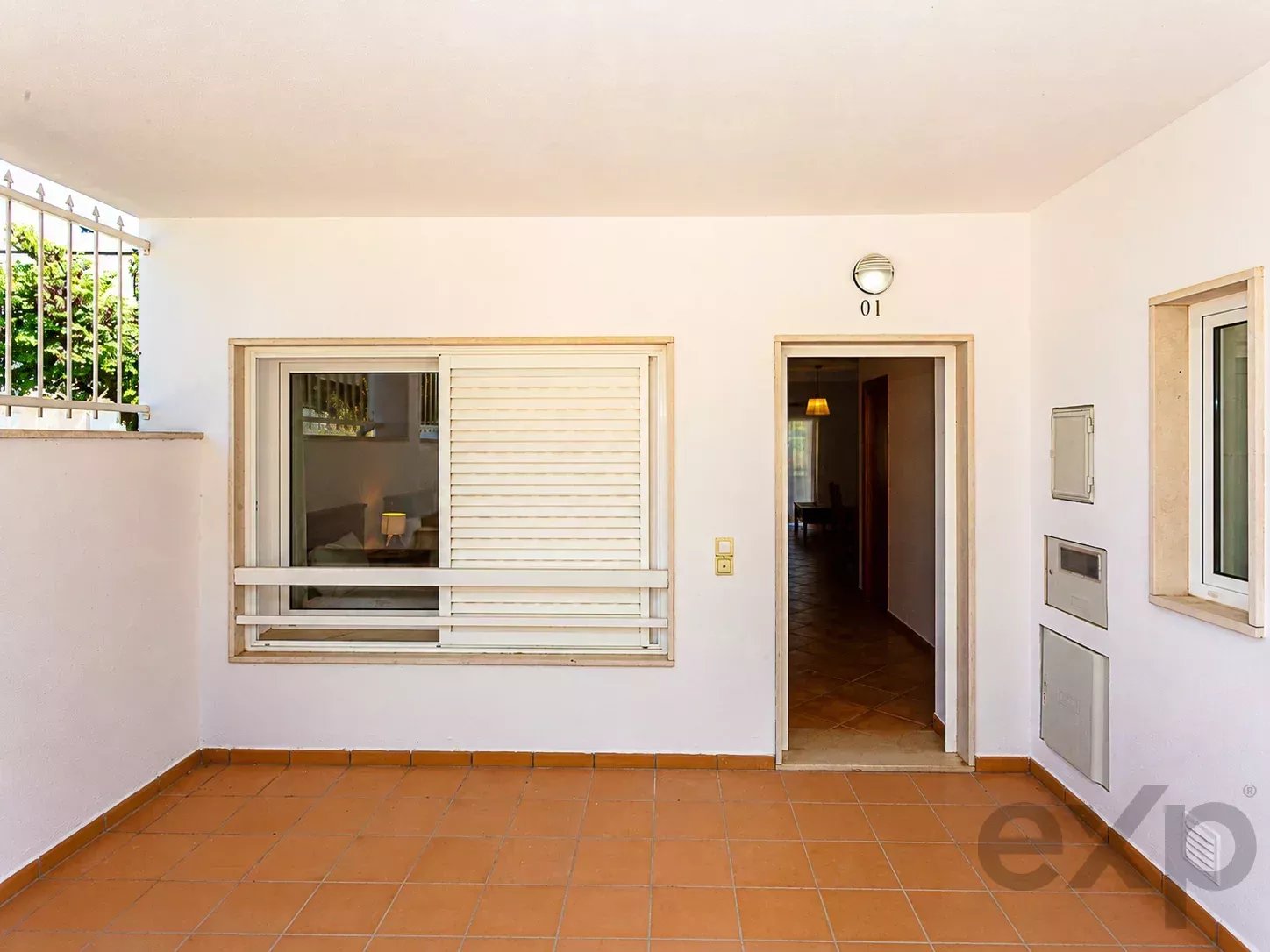 Covered apartment entrance: white walls, terracotta tile floor, a window with shutters on the left, and a doorway labeled 01 opening into a dimly lit hallway; mailbox panels are on the right wall.