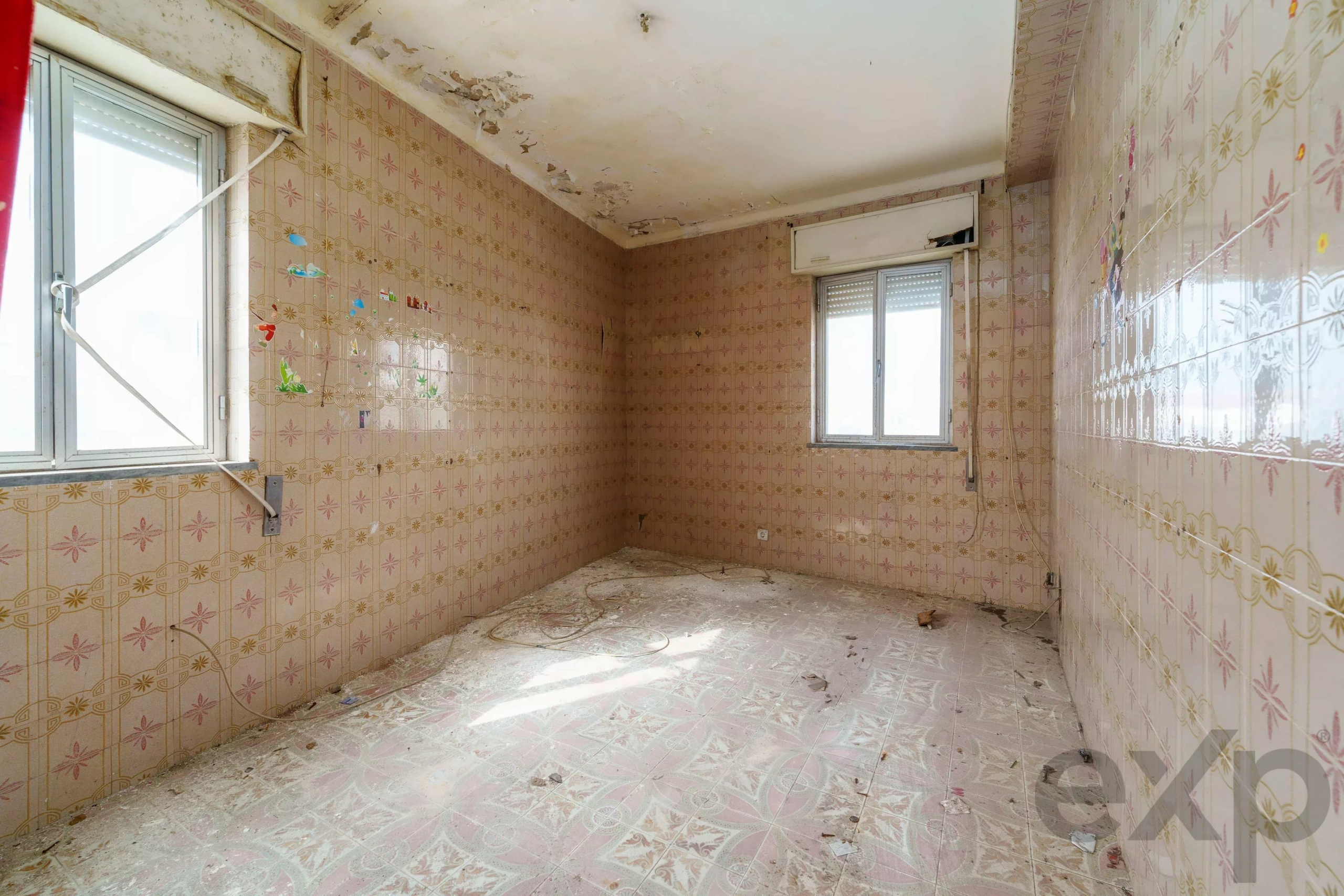 Abandoned room with pink patterned tiles, peeling ceiling, two windows, and scattered debris on the dusty floor.