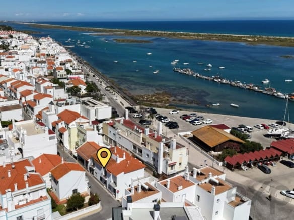 Aerial view of a coastal town with white buildings and orange roofs along a harbor and blue sea beyond; a yellow location pin marks a building in the foreground.