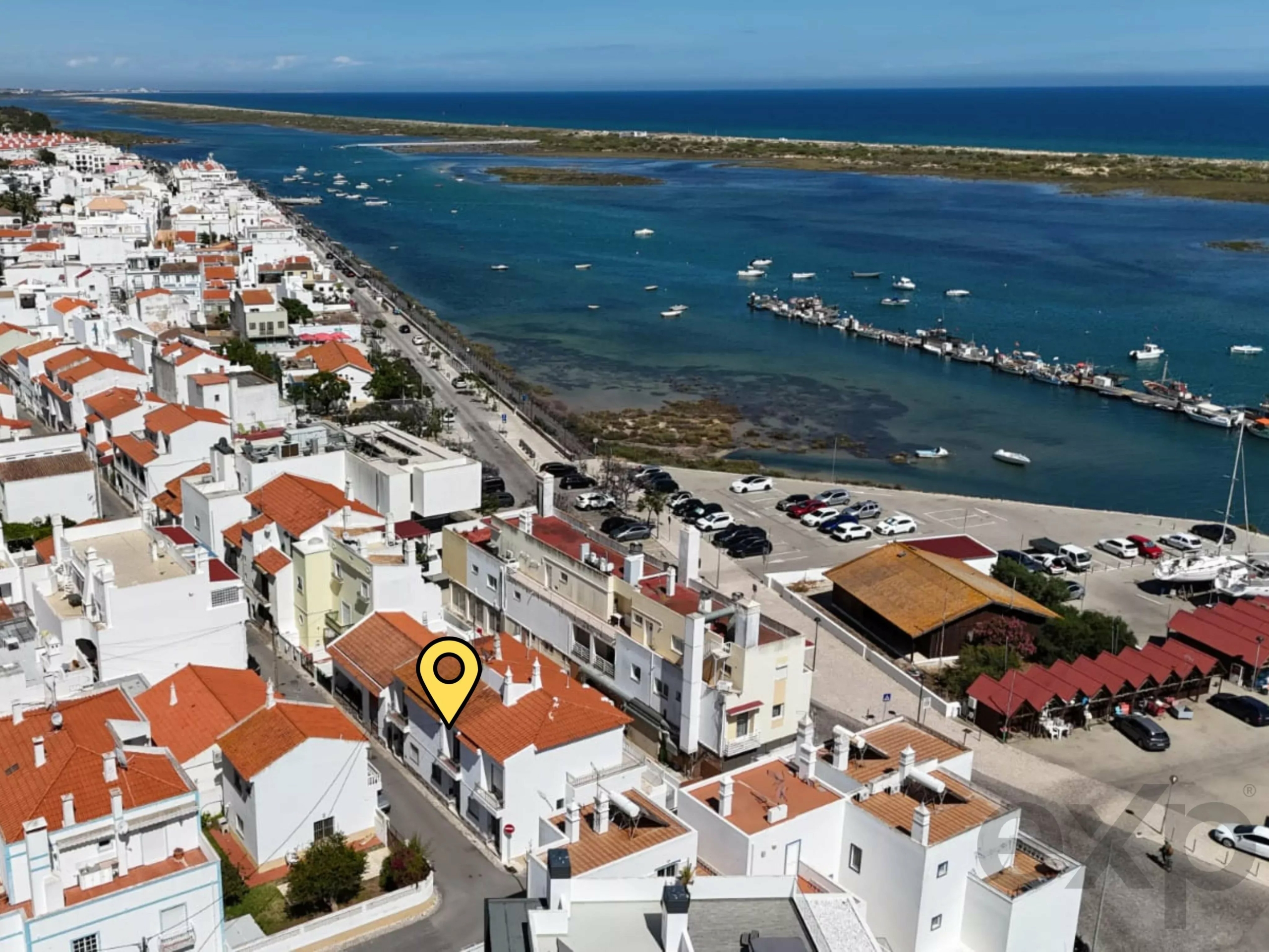 Aerial view of a coastal town with white buildings and orange roofs along a harbor and blue sea beyond; a yellow location pin marks a building in the foreground.
