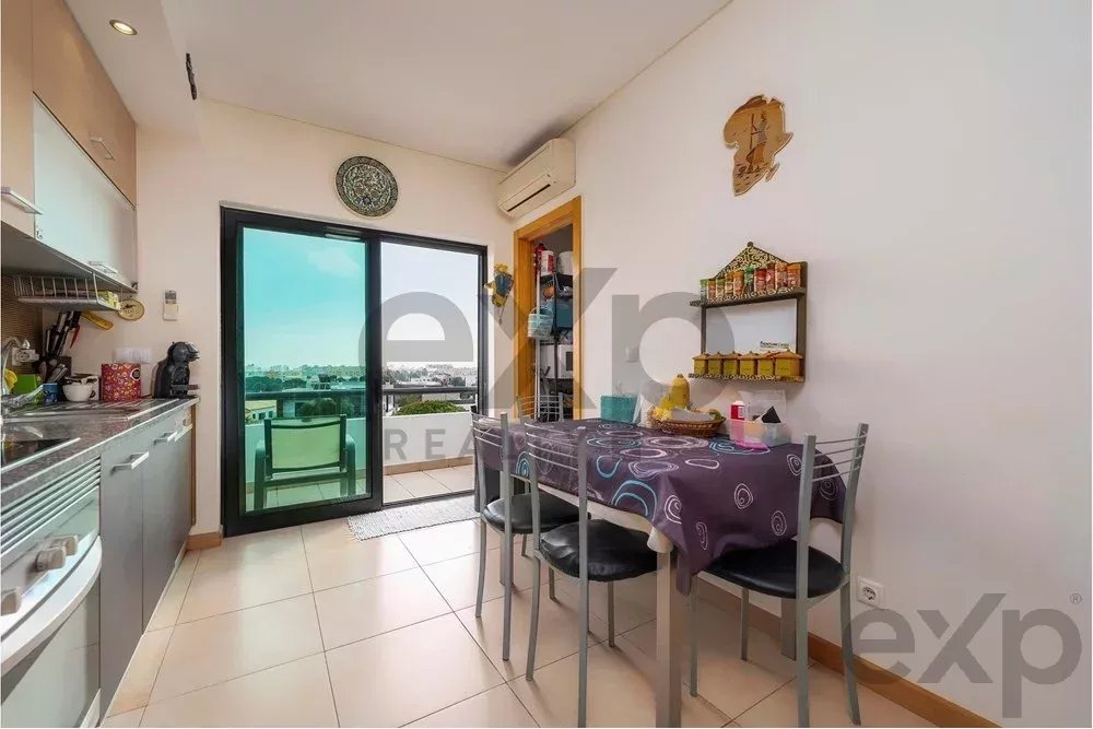 Compact kitchen with a four-seat dining table, leading to a balcony with city view. Aisle of grey cabinets and appliances along the left wall.