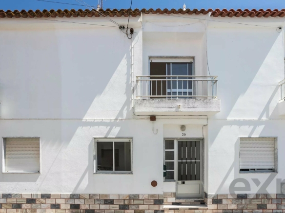 White building facade with a small balcony over a door, two windows with shutters, and a tiled base along the ground.