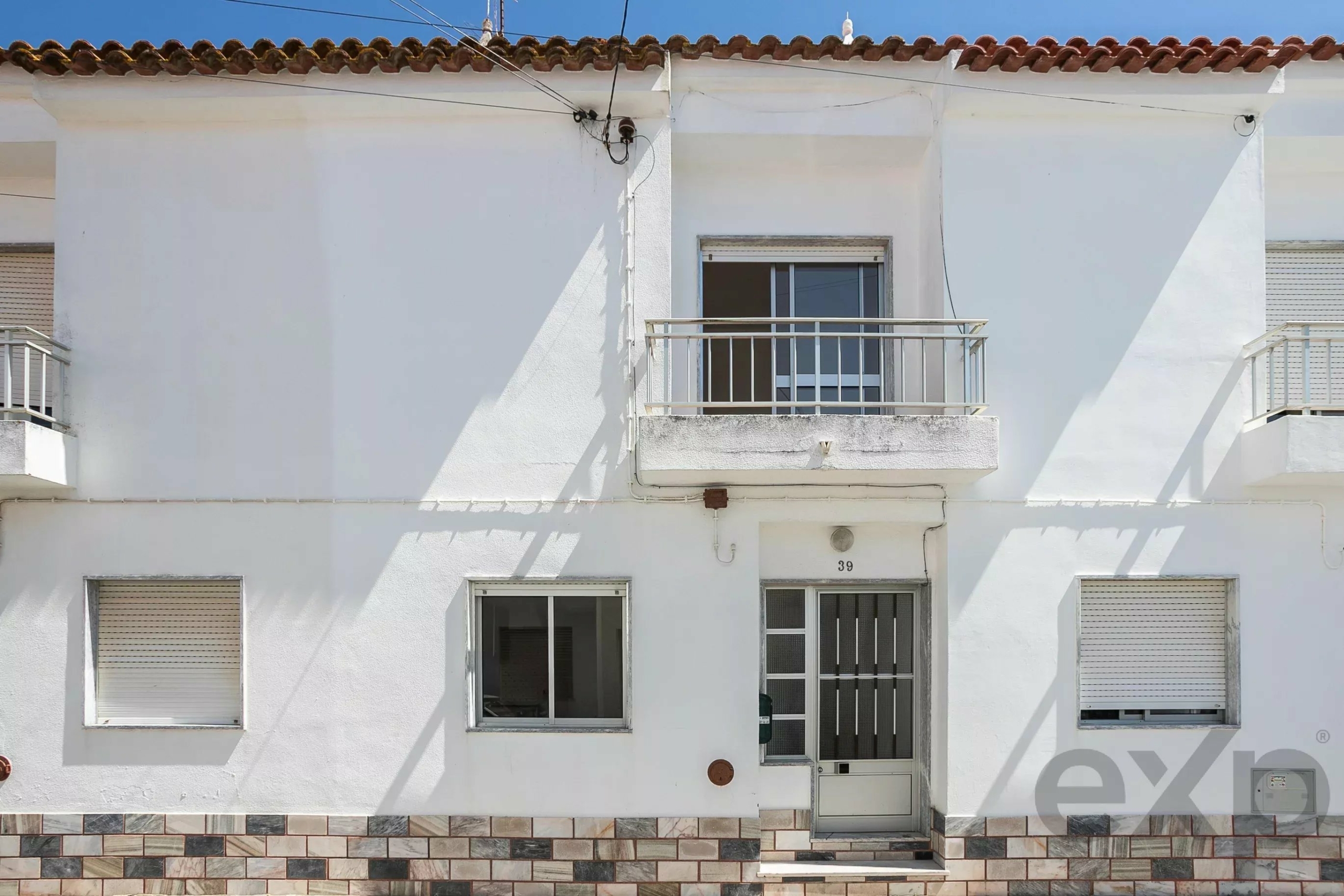 White building facade with a small balcony over a door, two windows with shutters, and a tiled base along the ground.