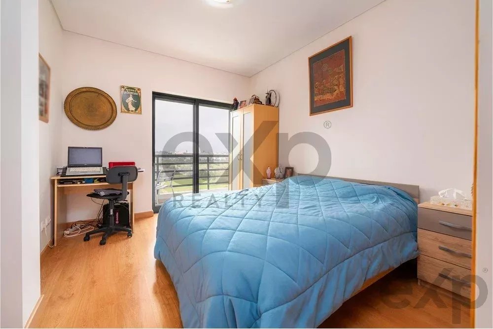 Bedroom with a blue quilted bed, desk with a laptop, and a large window/ balcony door nearby.