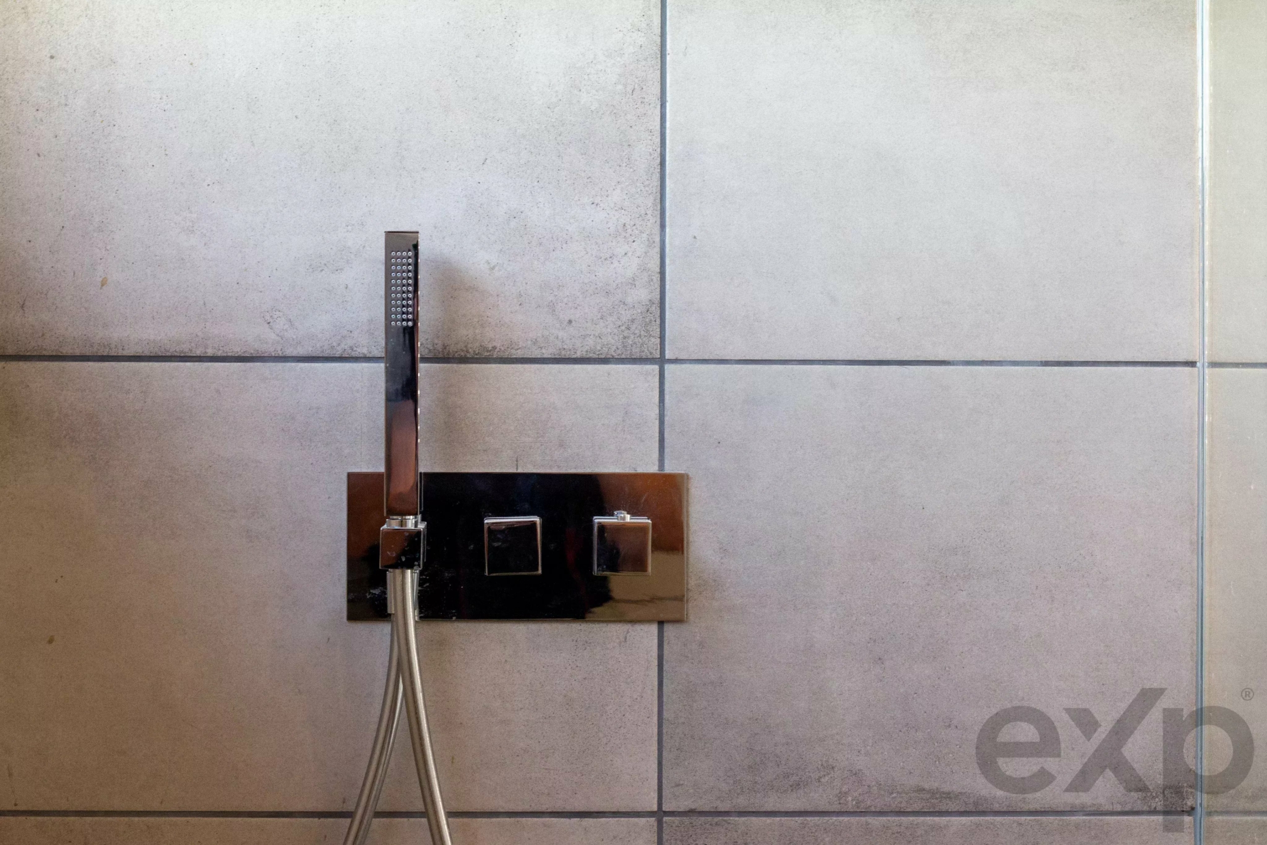 Chrome rectangular shower fixture with vertical handheld shower head mounted on a gray tiled bathroom wall.