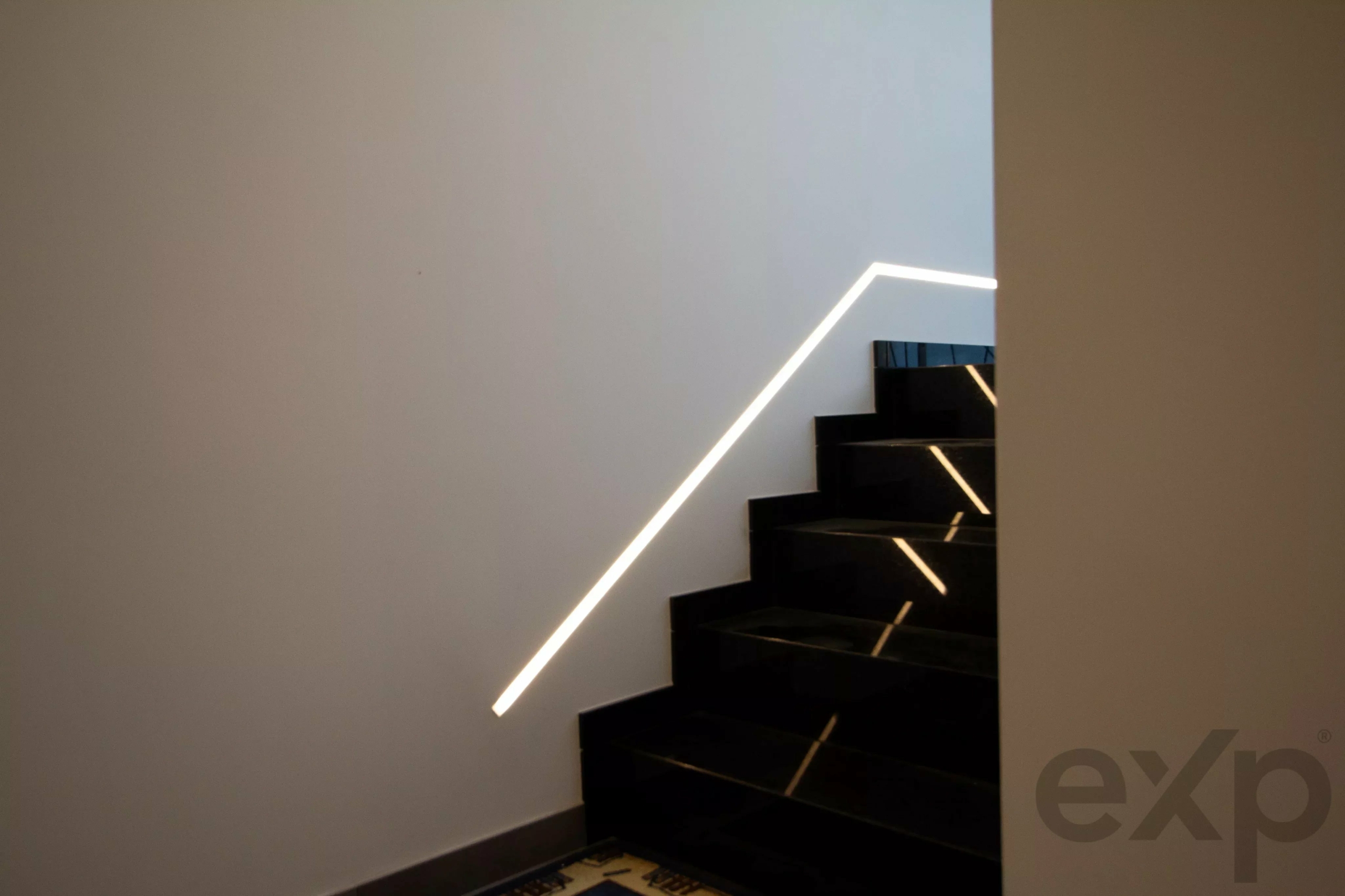 Modern staircase with a glowing white handrail along a white wall; dark steps and a partial logo visible in the lower-right corner.