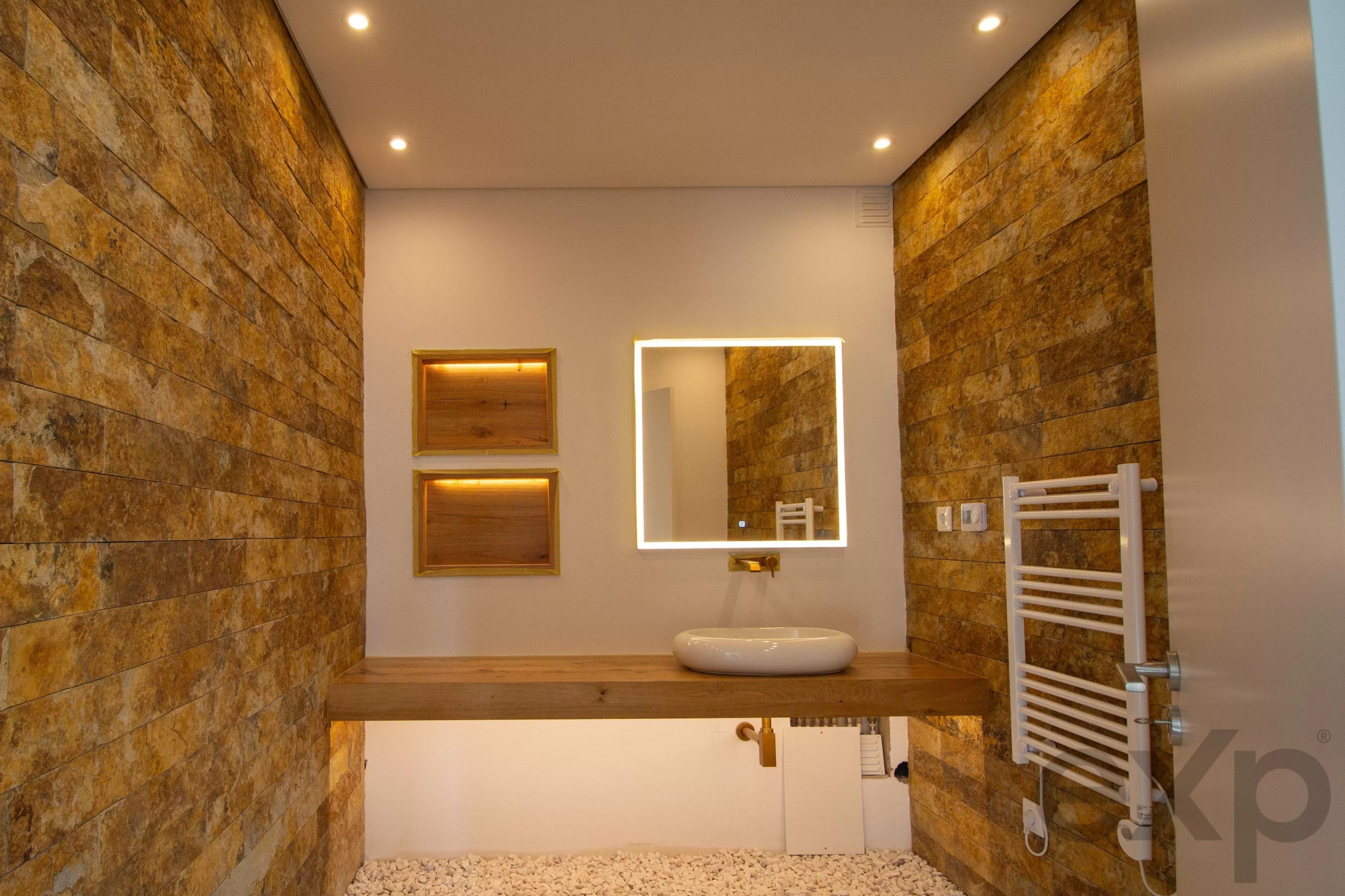 Modern bathroom with warm stone-tiled walls, a backlit square mirror above a white vessel sink, and wooden shelves on the wall.