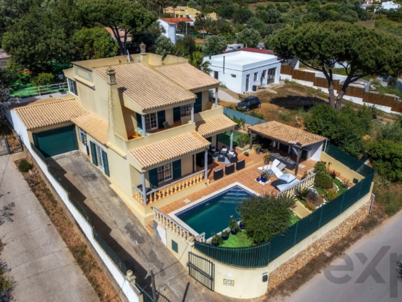 Aerial view of a yellow Mediterranean villa with tiled roof, swimming pool, and patio surrounded by trees and other houses.