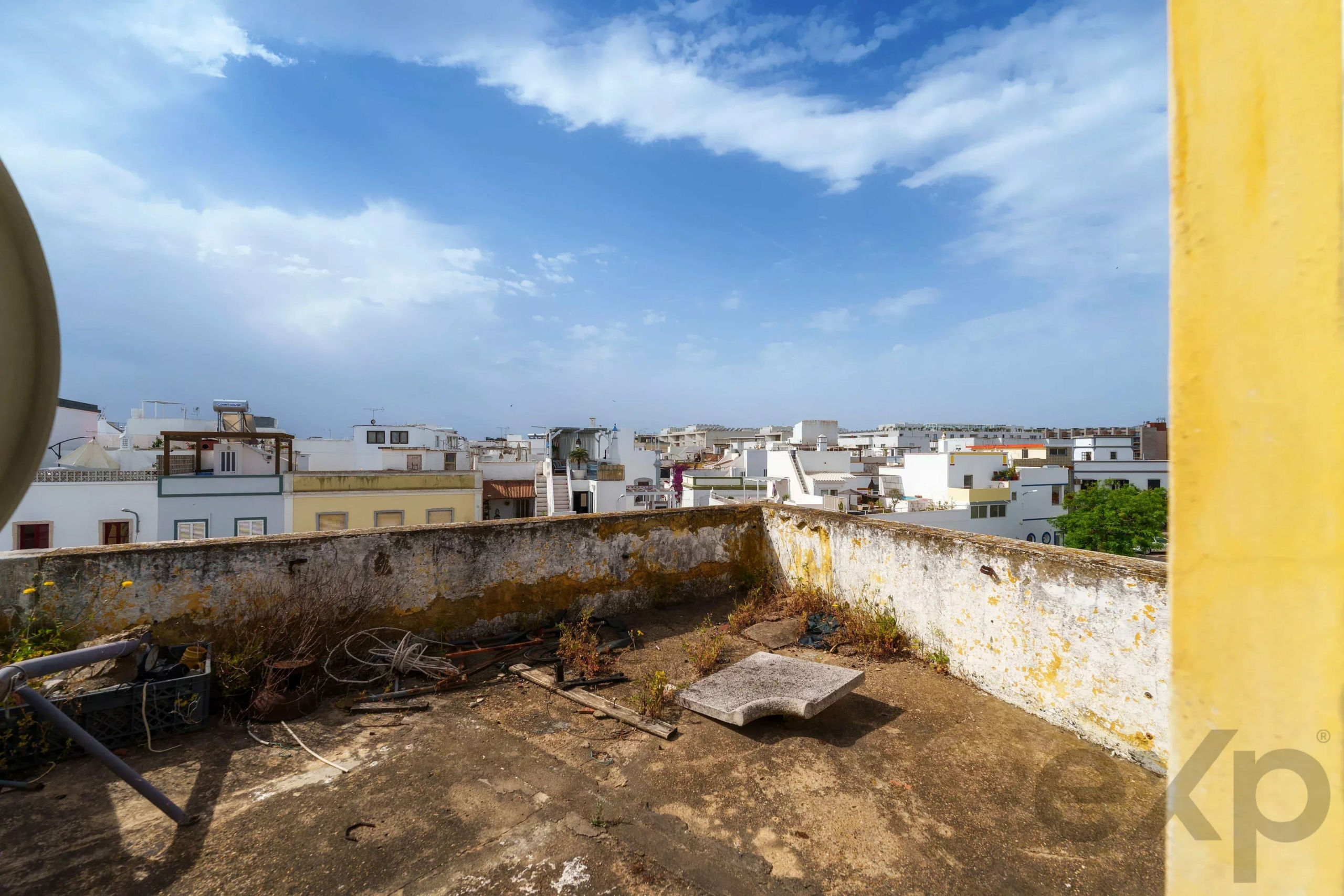 Rooftop area with rubble, pipes, and weeds, overlooking white buildings under a clear blue sky with a yellow pillar on the right edge nearby.