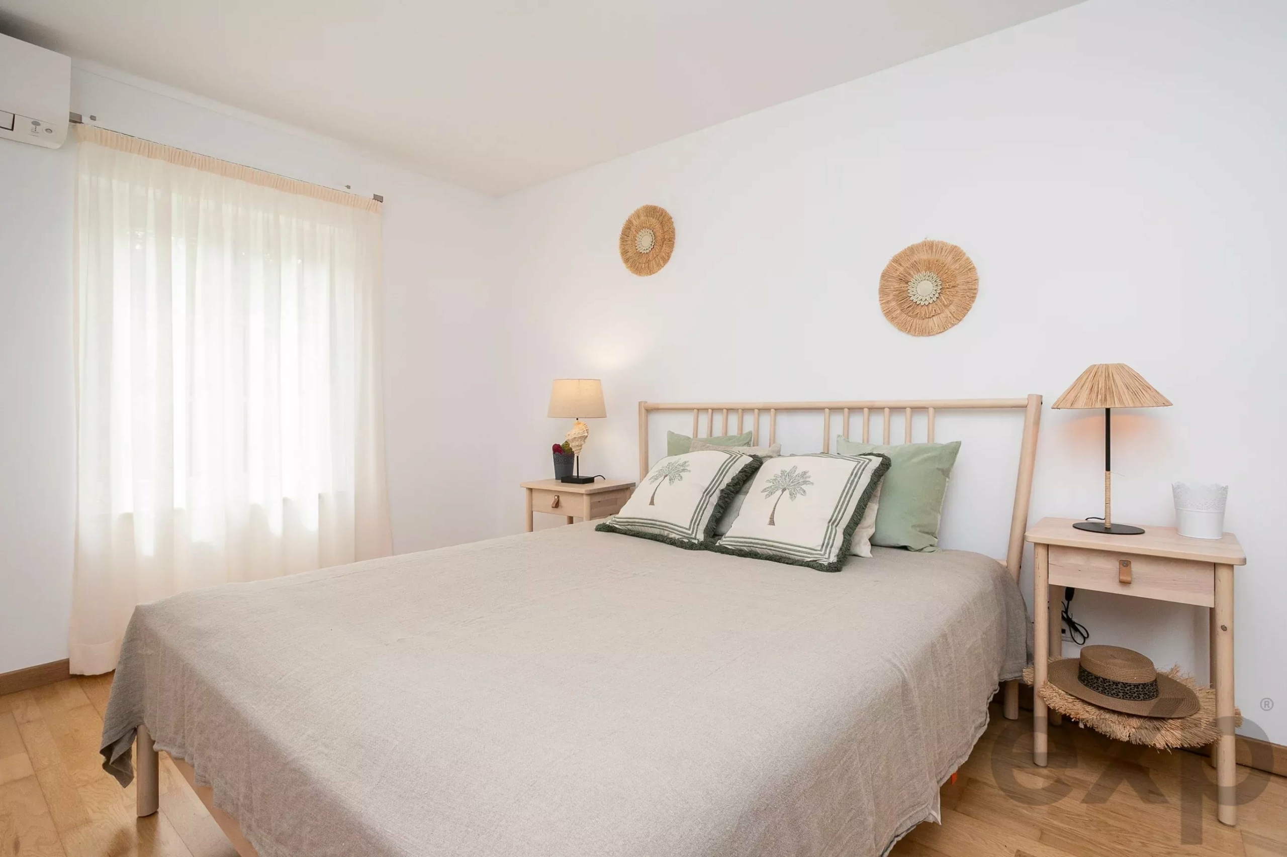 Bright bedroom with a beige bedspread, wicker wall decor, two nightstands, and sheer curtains on the left.