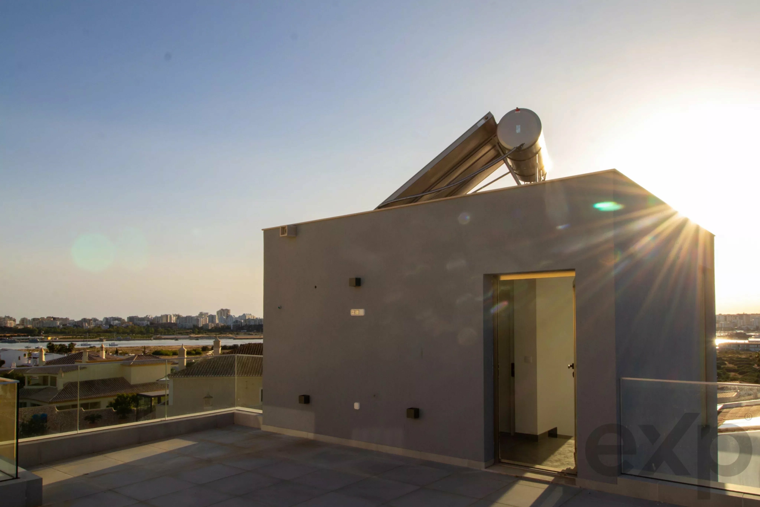 Rooftop terrace with a circular solar water heater on a tilted frame and a distant city skyline at sunset.
