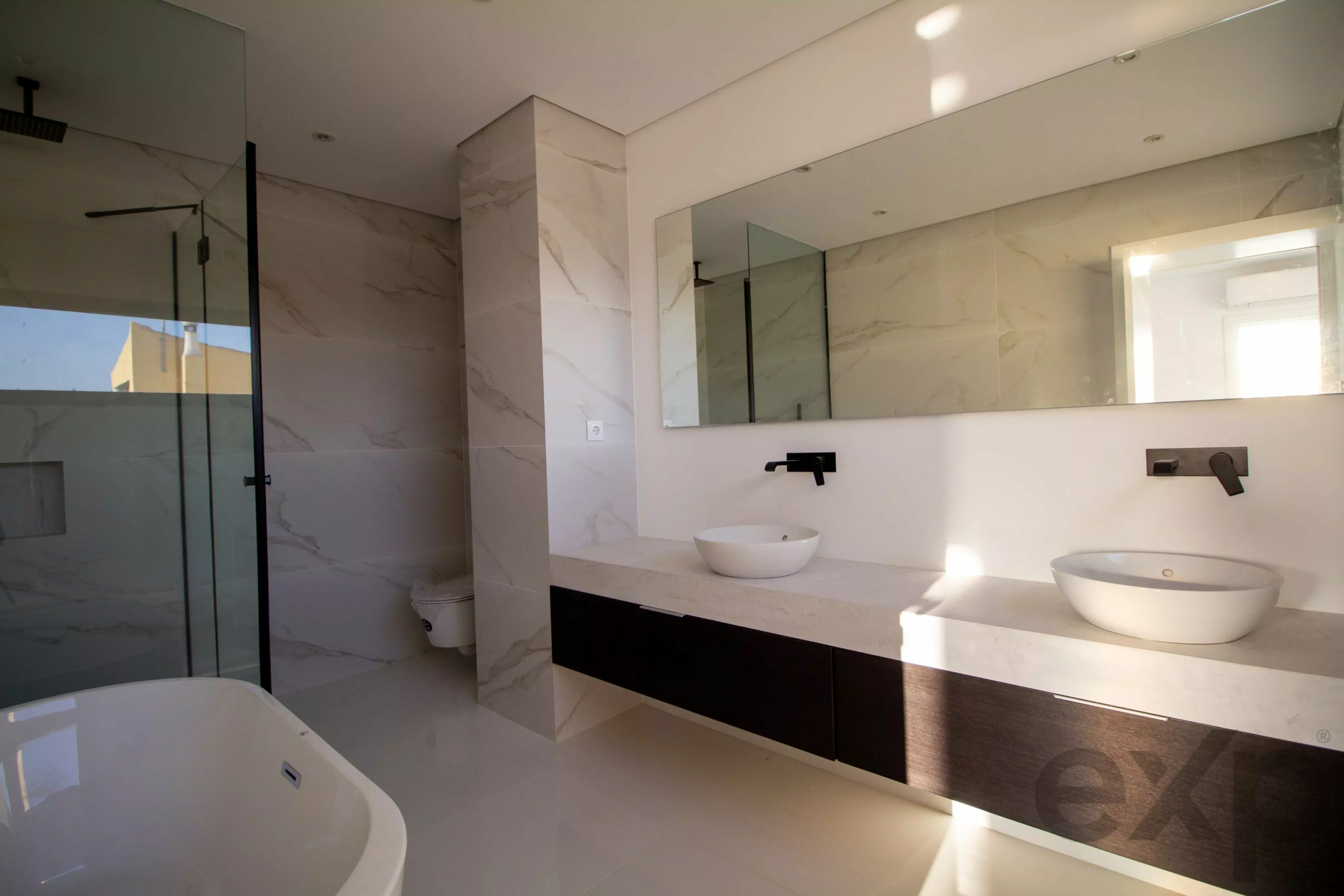 Modern bathroom with white marble walls, a long countertop, and two round vessel sinks beneath a wide mirror.