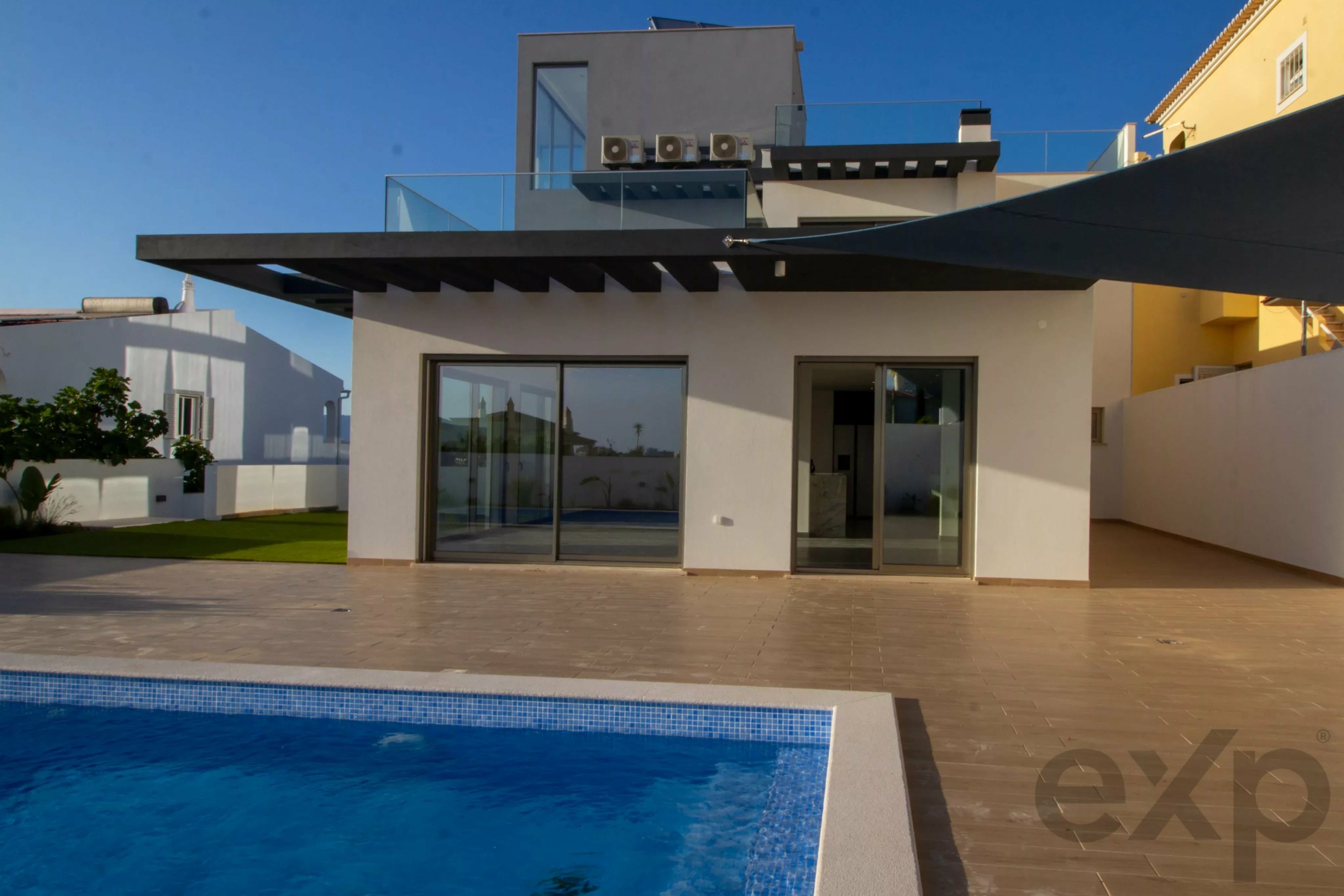 Modern two-story house with large glass doors and a rooftop terrace, overlooking a blue-tiled swimming pool in the foreground. The patio is beige-tiled with a dark overhang and open-plan living space visible inside.