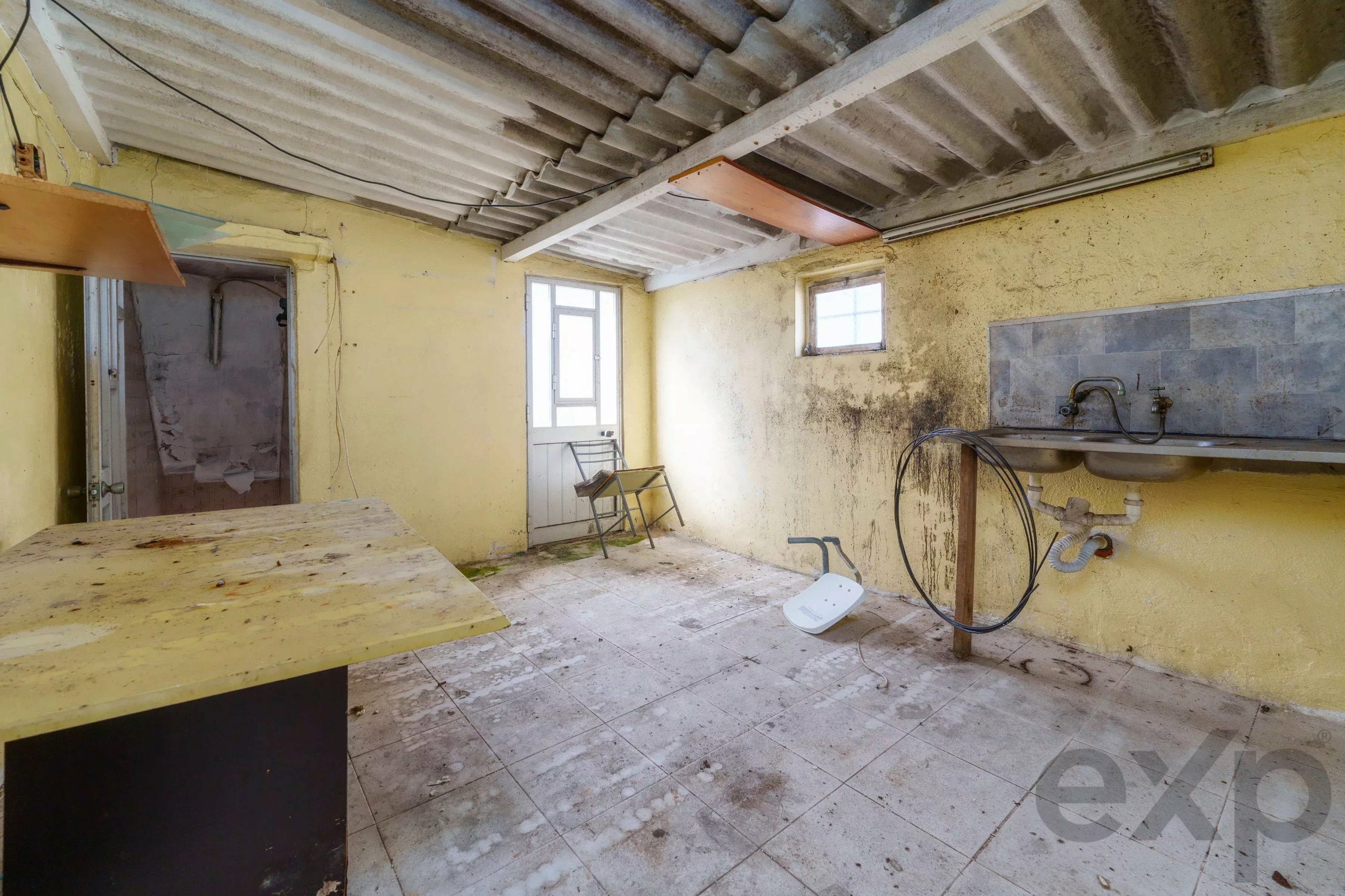 Abandoned yellow kitchen with a dirty sink, exposed pipes, and a corrugated ceiling; sunlight from a small window reveals peeling walls and grime.