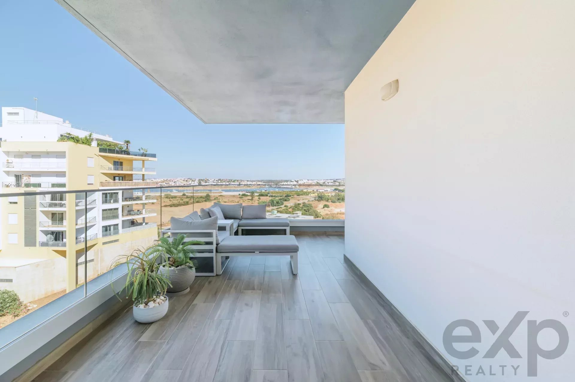 Modern balcony with gray outdoor seating, potted plants, and a glass railing overlooking a coastal cityscape.