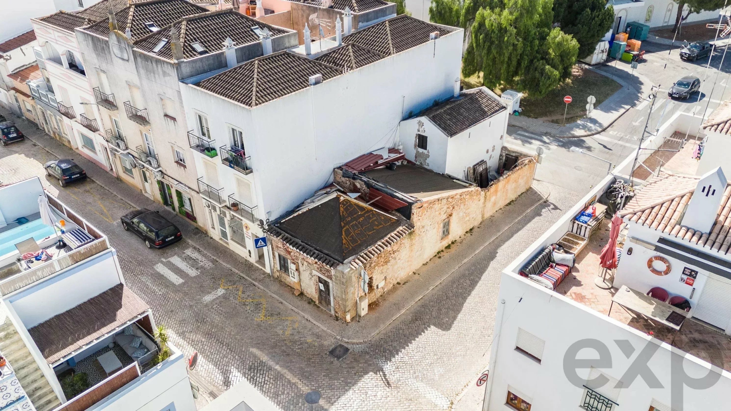 Aerial view of a narrow cobblestone street lined with white townhouses, tiled roofs, balconies, and parked cars in a coastal alleyway.