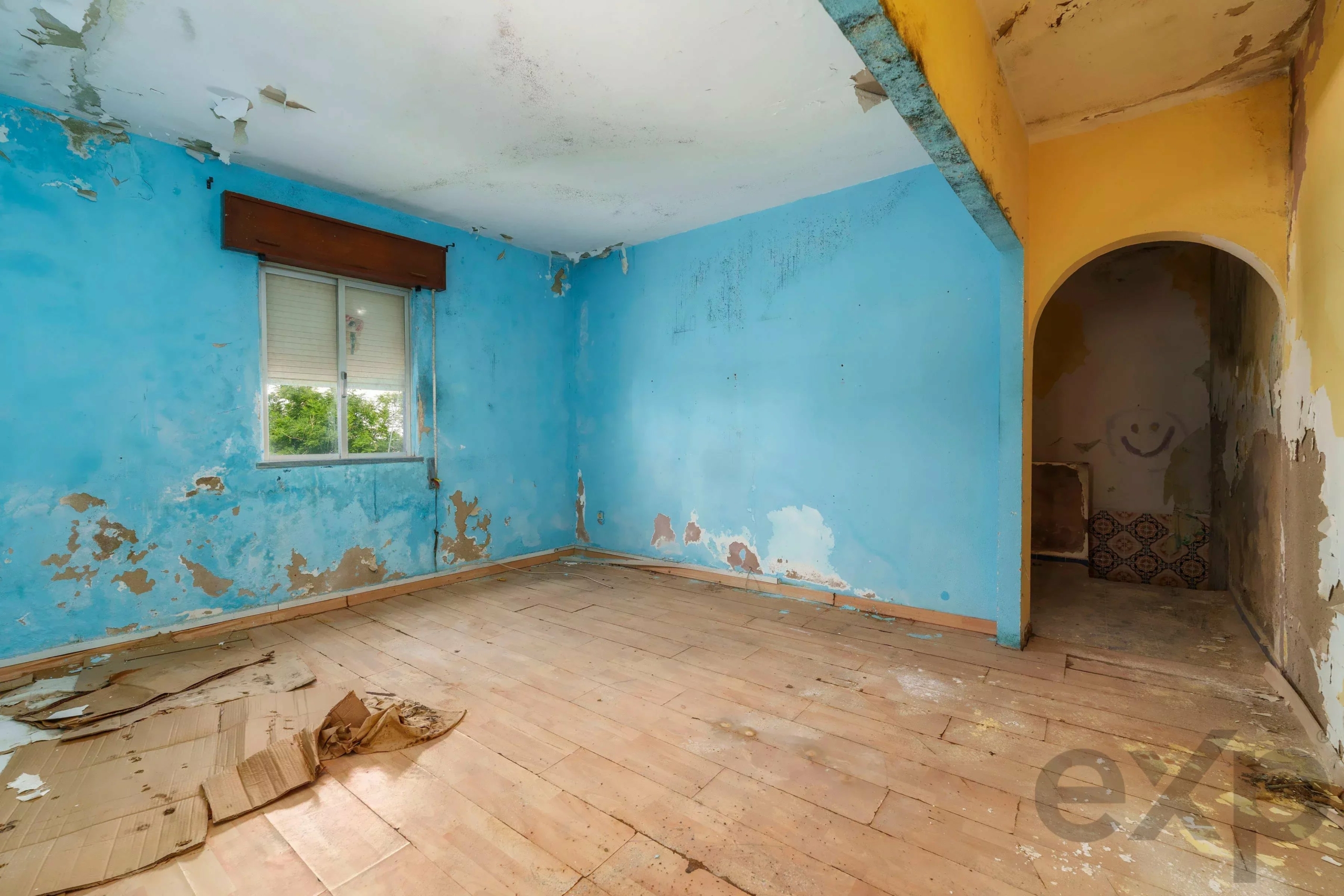 Abandoned blue-walled room with peeling paint, scattered debris on the wooden floor, and a yellow archway leading to a dark doorway.