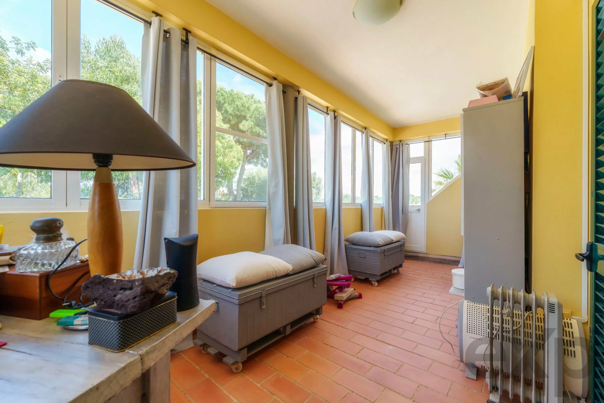 Bright sunroom with yellow walls, large windows, and gray curtains; benches with cushions along the windowed wall and a table with a lamp and decorative stones in the foreground.