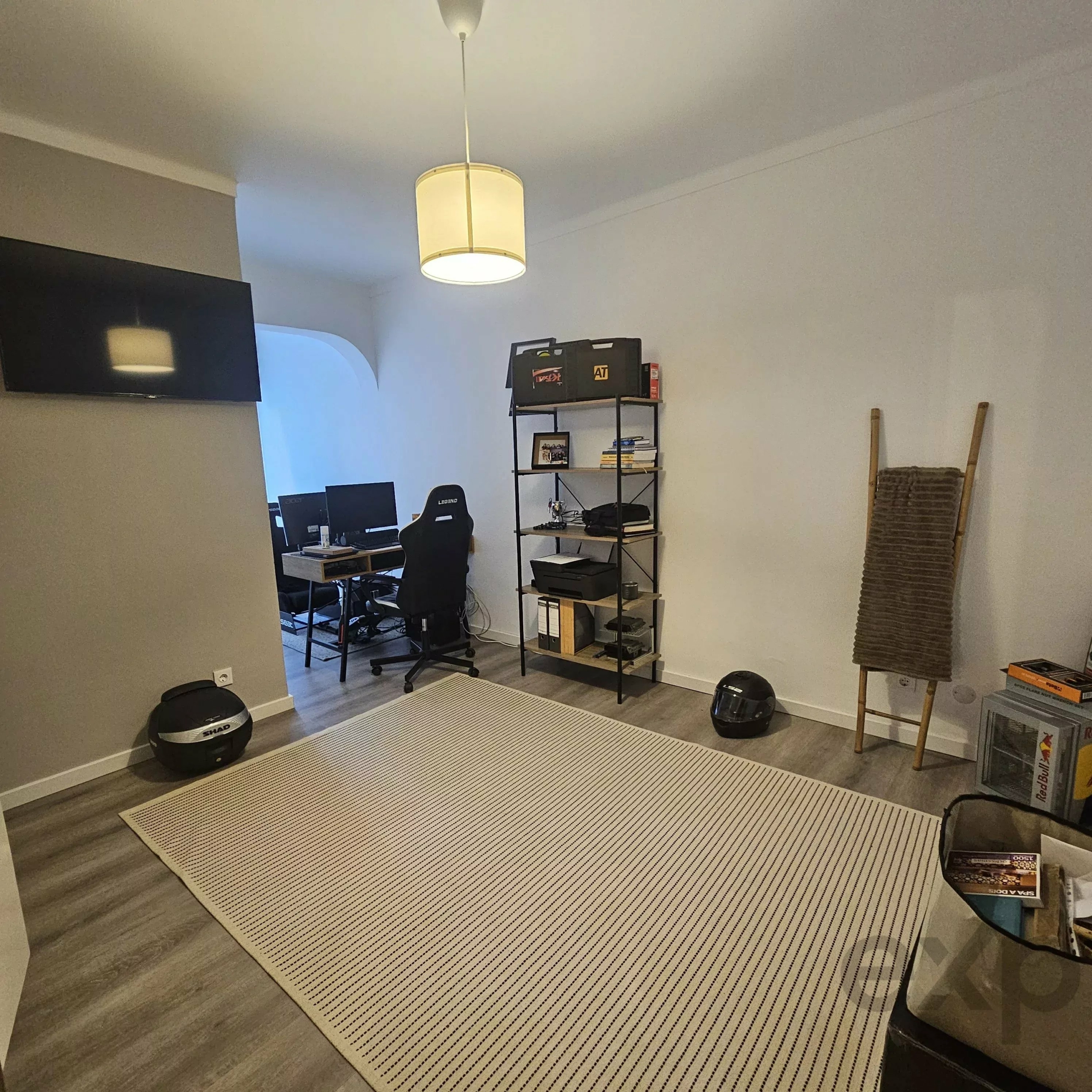 Home office with a striped rug, desk and monitors on the left, shelves and boxes in the back, and a woven chair on the right corner.