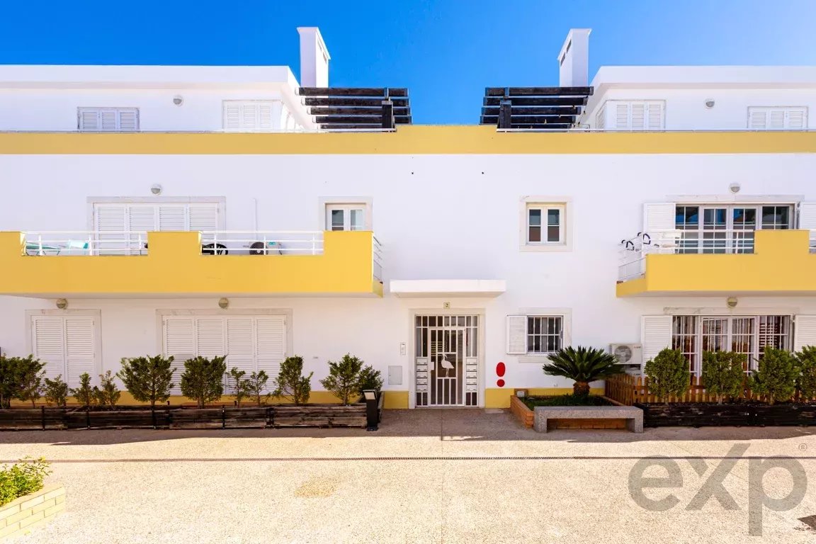White apartment building with bright yellow balconies under a blue sky, featuring a central entrance and shrubs along the front.