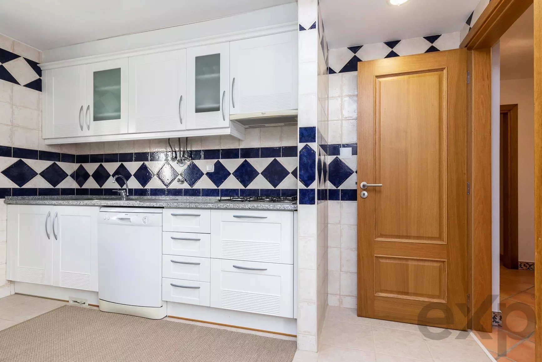 White kitchen with upper and lower cabinets, blue-and-white diamond tile backsplash, and an open wooden door to a hallway on the right.
