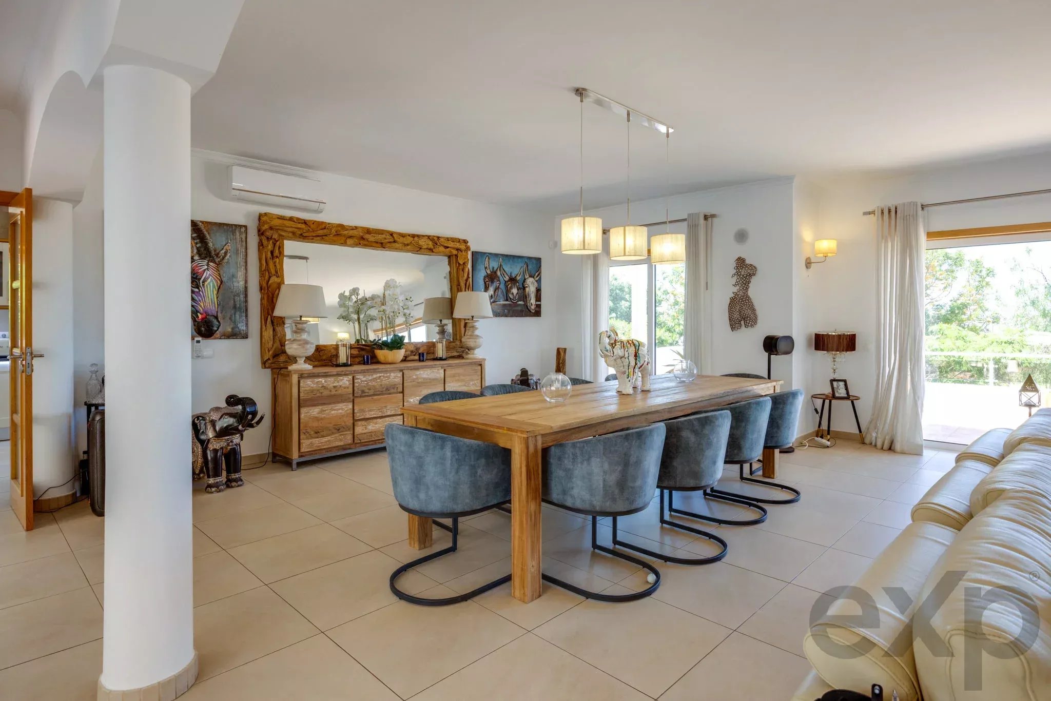 Bright open-plan dining area with a wooden table, blue upholstered chairs, and a large rustic mirror reflecting plants and lamps. The room is sunlit by large windows.