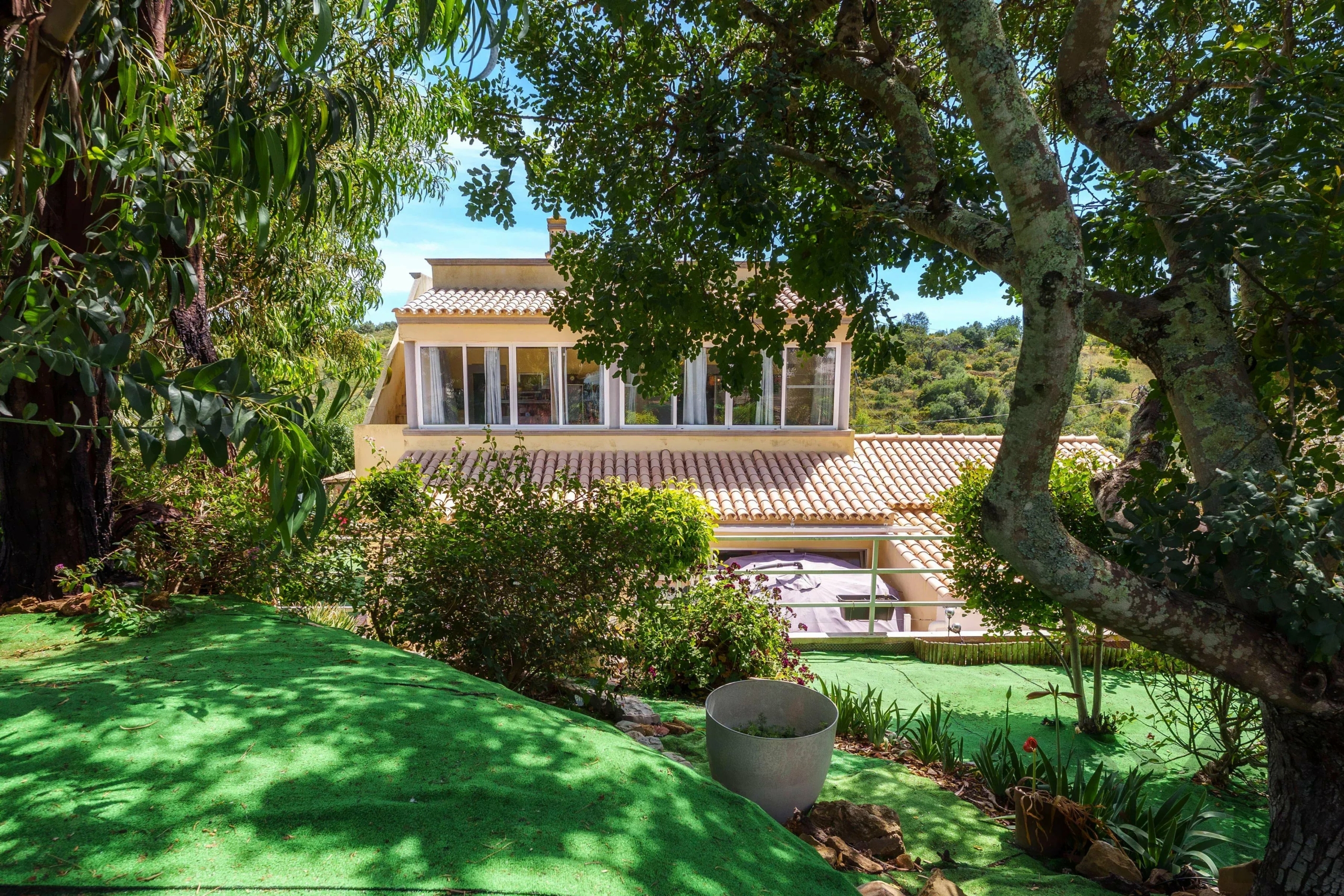 House with terracotta-tiled roof seen through dense green trees in a sunny garden courtyard; foreground is green lawn and potted plants.