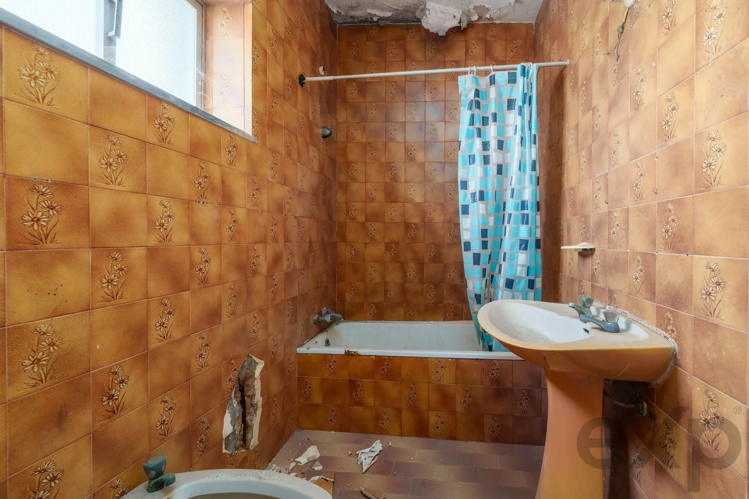 Dilapidated orange-tiled bathroom with a blue patterned shower curtain and a white sink, showing damaged ceiling and debris on the floor.