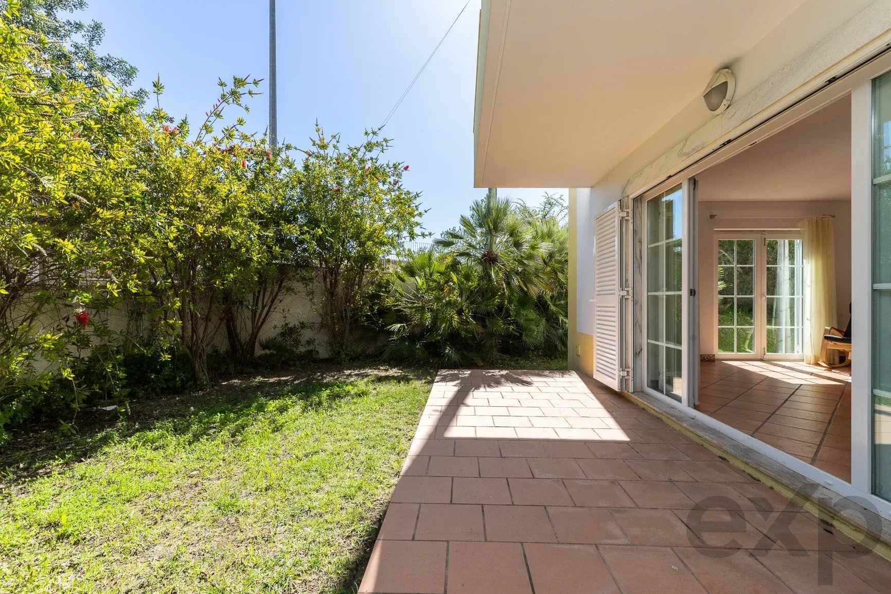 Patio beside a house with sliding glass doors open to a sunlit living area; dense green shrubs along the boundary left.