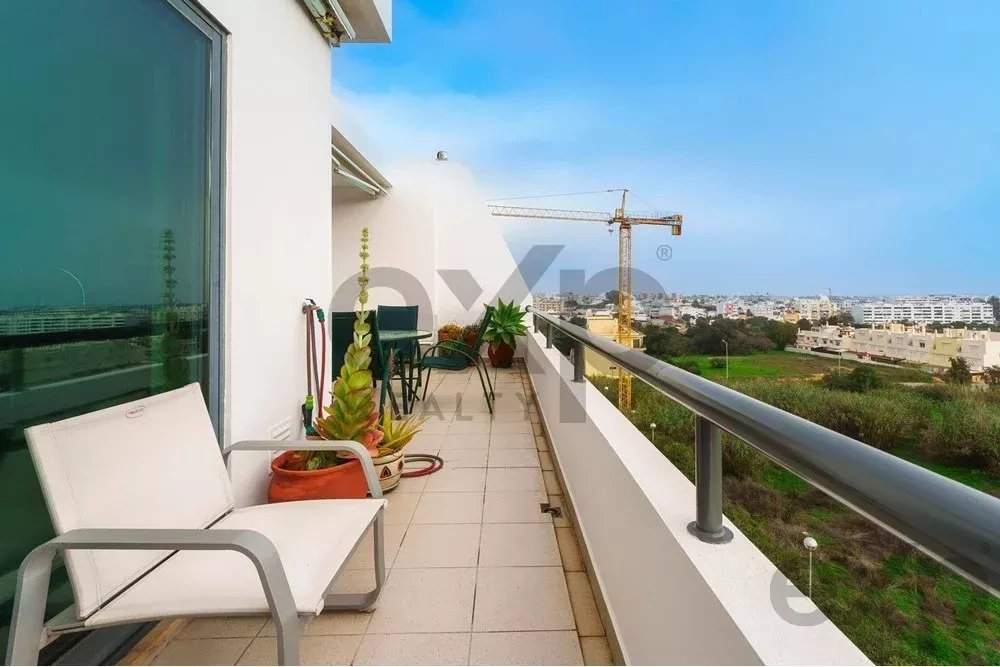 Balcony with white chairs and potted plants overlooking a residential area and a construction crane in the distance.