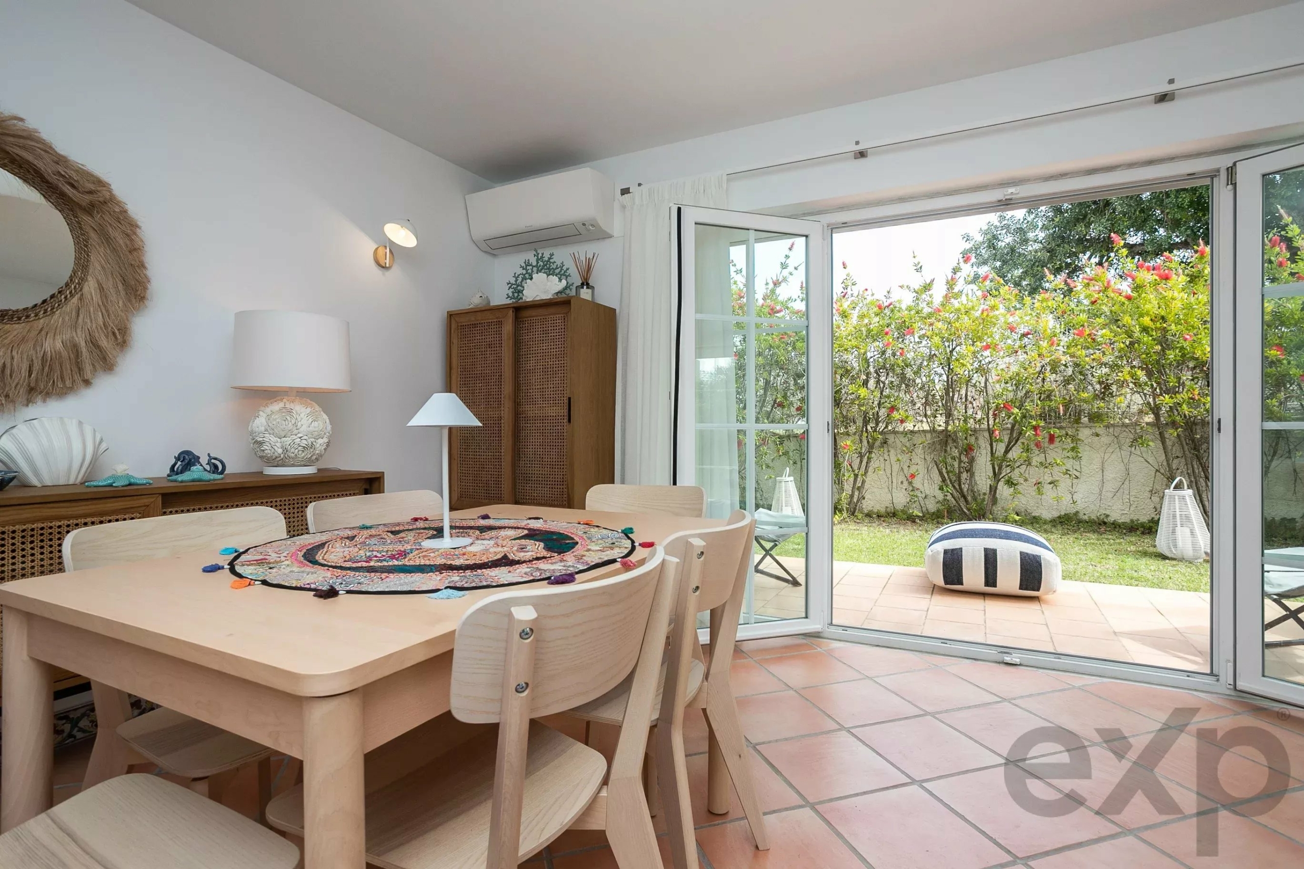 Bright dining area with a wooden table and chairs, open glass doors to a garden outside. On the wall: wicker sunburst and sea-themed decor.