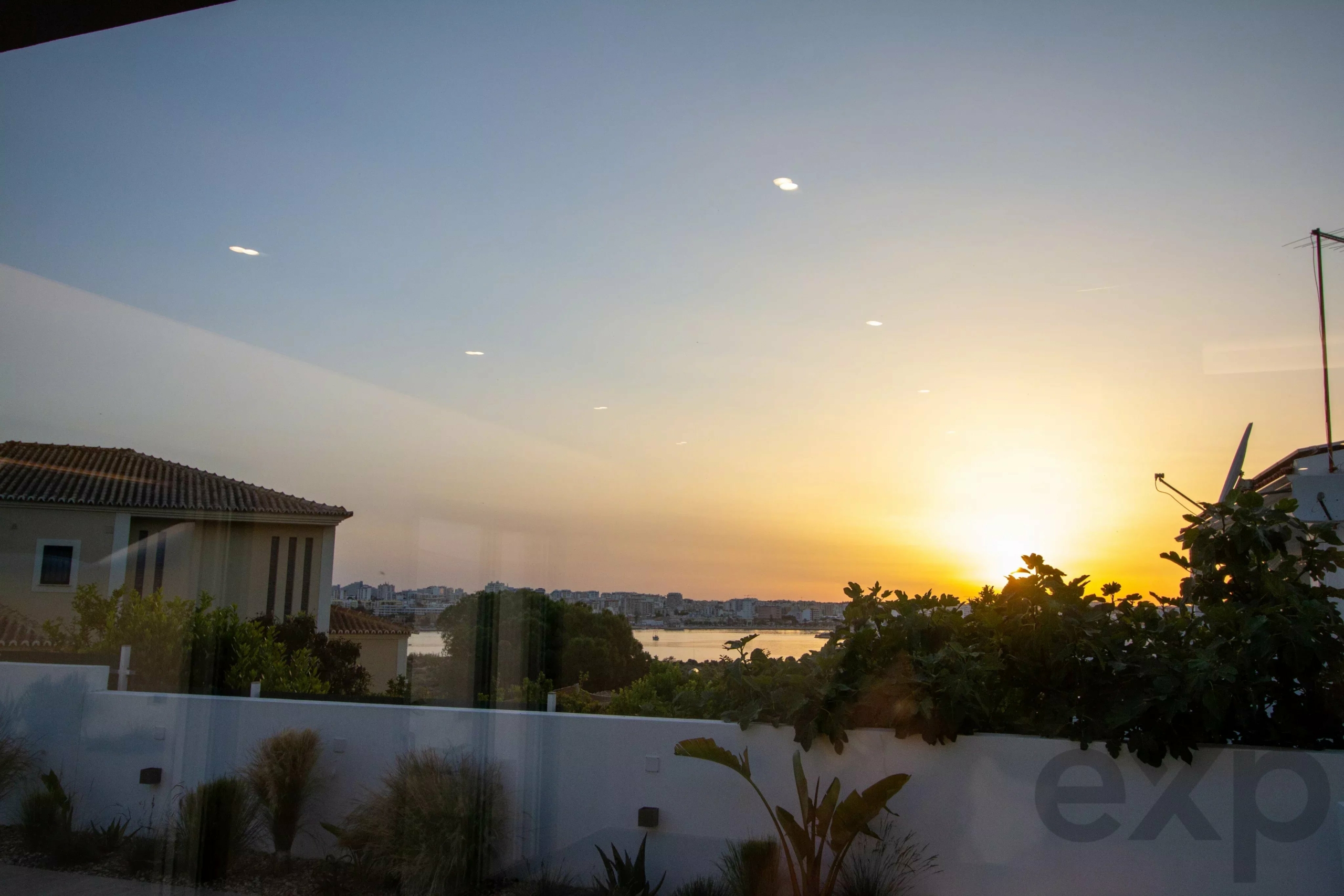 Sunset over a city skyline viewed through a glass balcony wall, with plants in the foreground and warm orange light near the horizon.