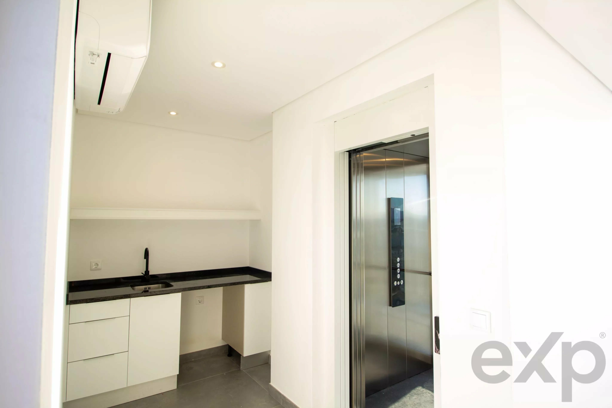 Small, modern kitchenette with white cabinetry, black countertop, and a sink on the left; an elevator door is on the right side of the image.