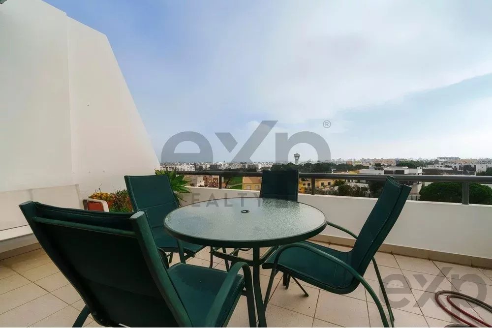 Rooftop balcony with a round glass table and four green chairs facing a city skyline under a blue sky.