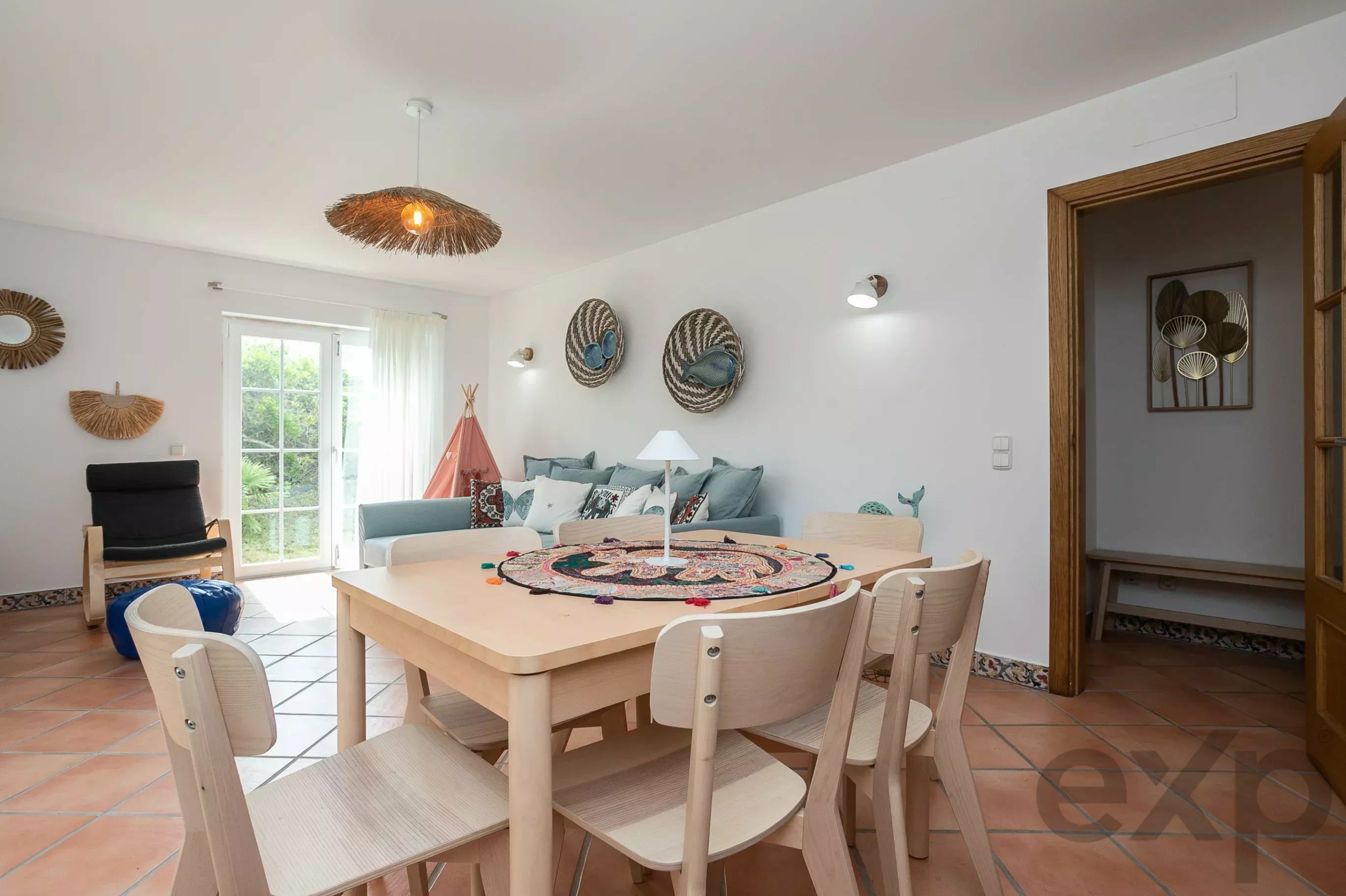 Bright dining room with a light wood table and chairs, round woven wall decor, and a sunburst pendant light overhead.