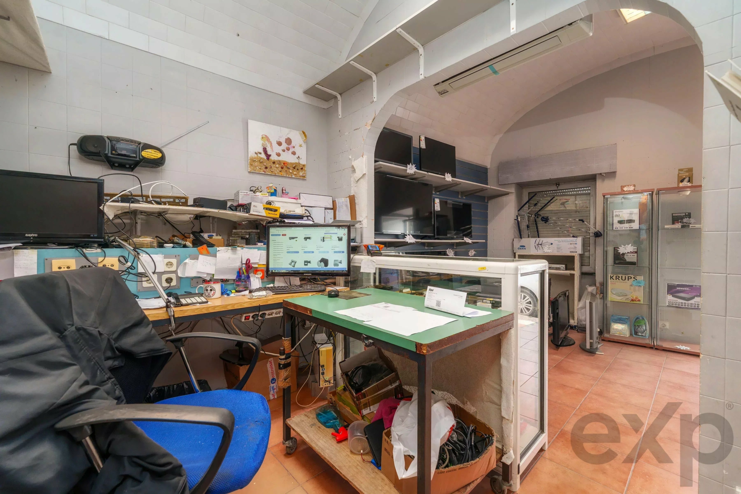 Cluttered electronics repair shop workspace with monitors, cables, and shelves of parts behind a glass display case.