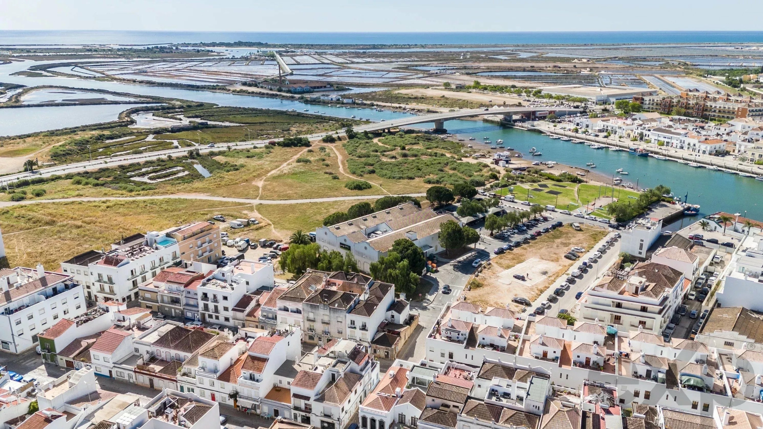 Aerial view of a coastal town with a canal network, bridge, boats, and white buildings along the waterfront near the sea.
