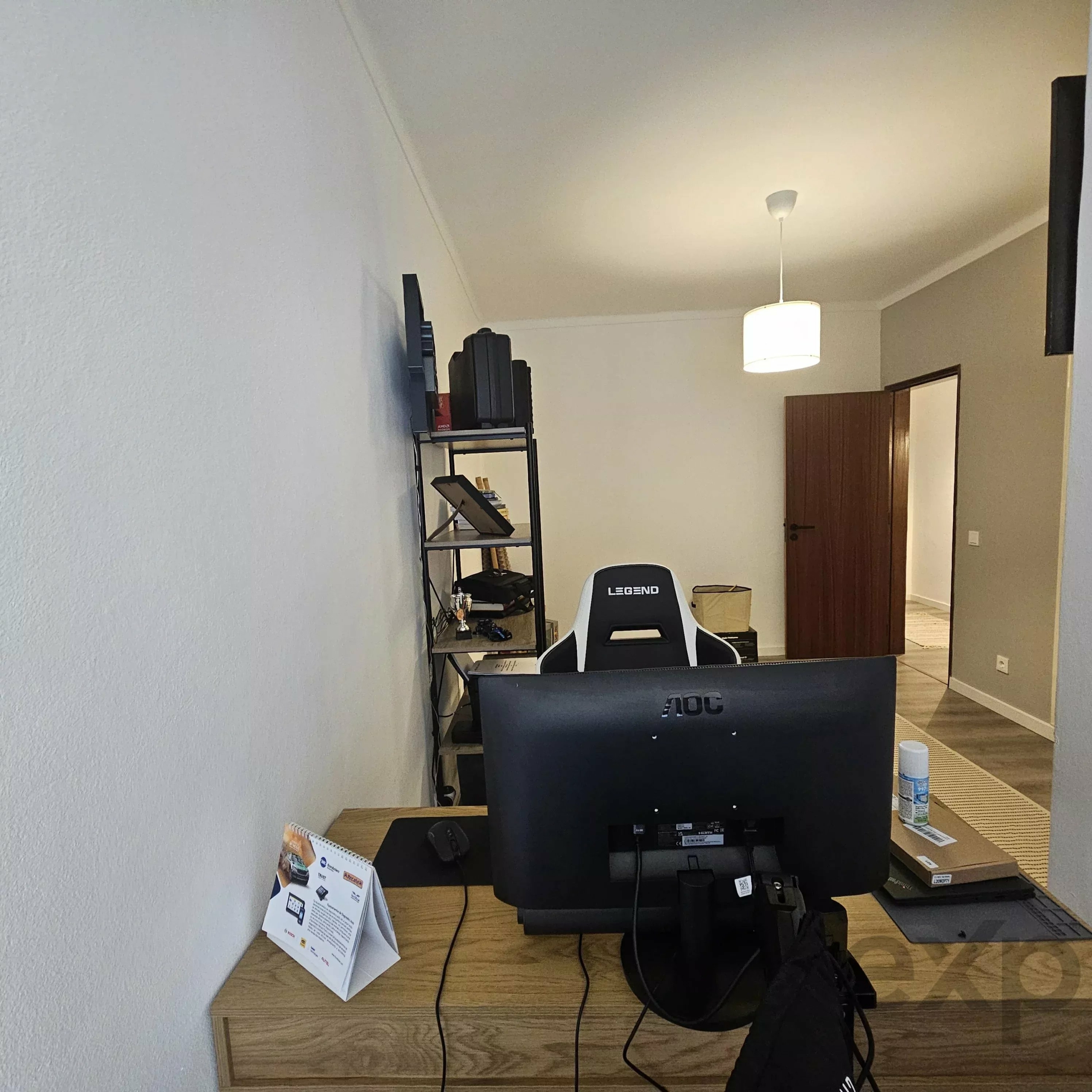 Office desk setup with a large monitor facing away from the wall, black shelving unit, and a white hanging ceiling light in a corner room.