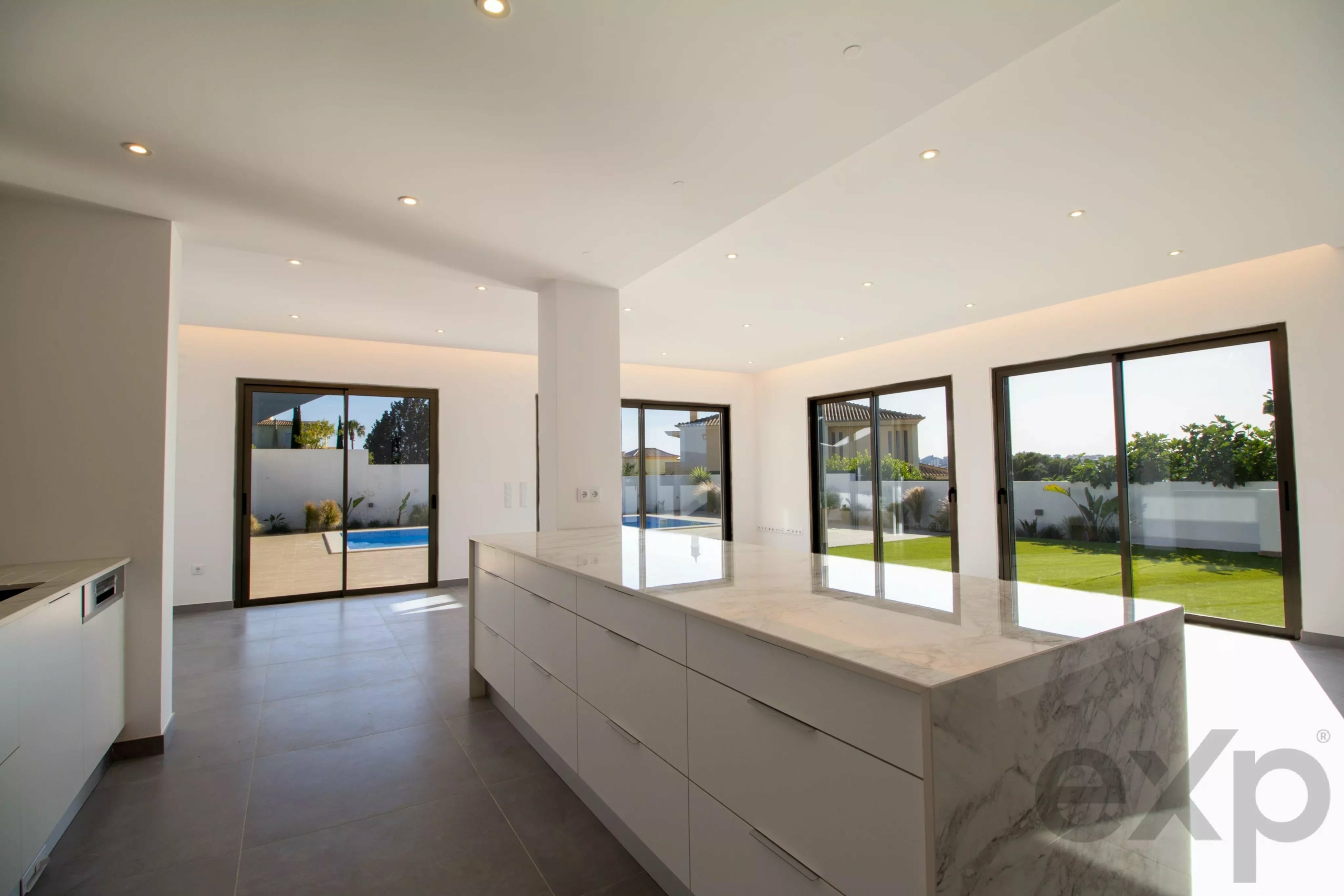 Bright modern kitchen with white cabinets and marble island, opening to a sunny backyard with a pool through large glass doors