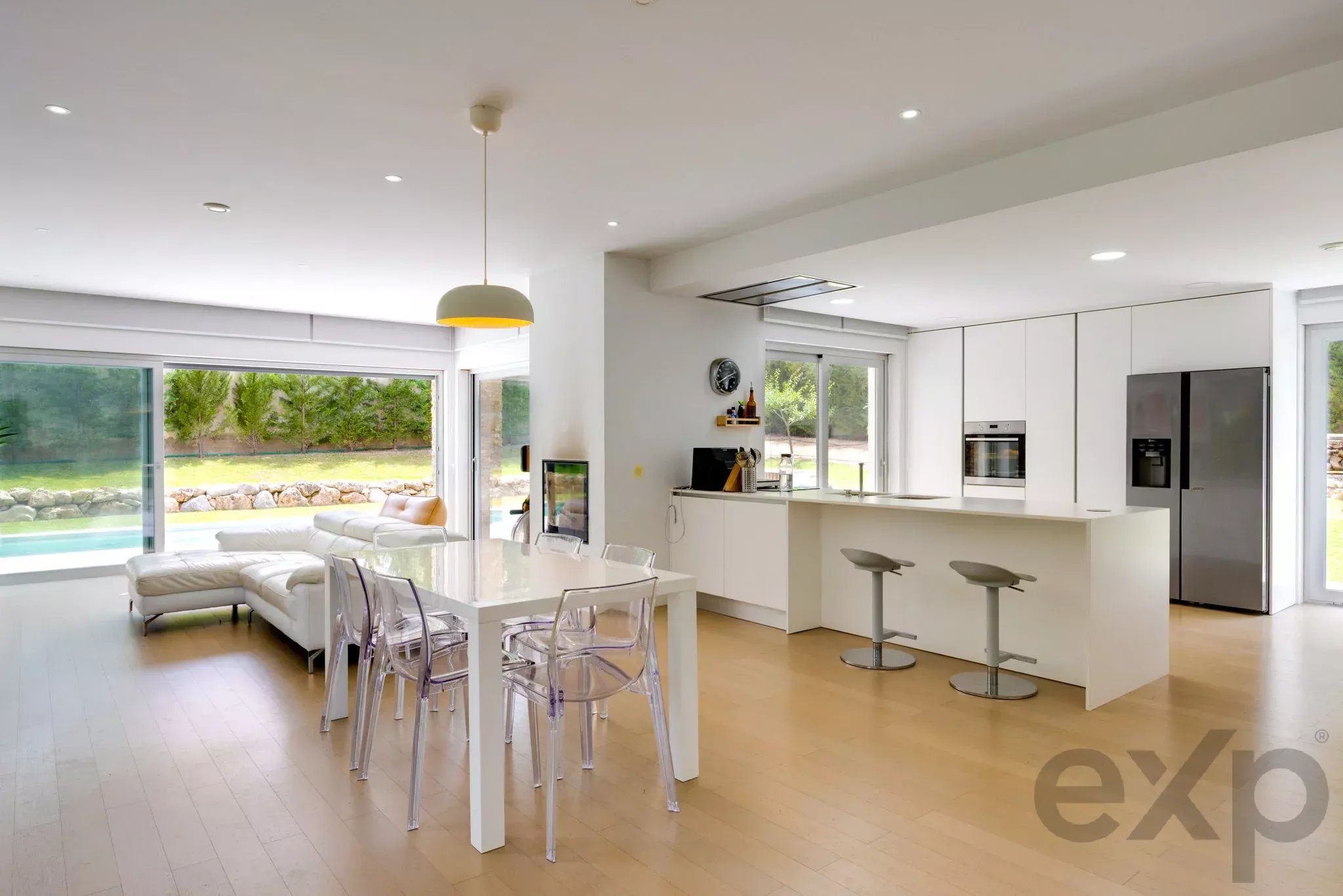 Bright open-plan kitchen and living room with white cabinets, island, glass chairs, and sliding doors to a green yard/pool area