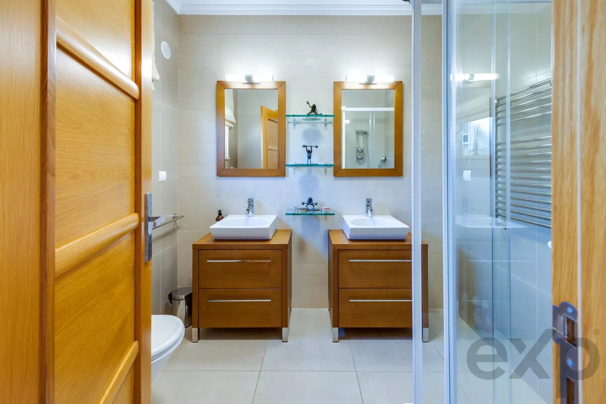 Modern bathroom with twin square sinks on wooden vanities, two framed mirrors, glass shelves, and a glass-enclosed shower to the right.