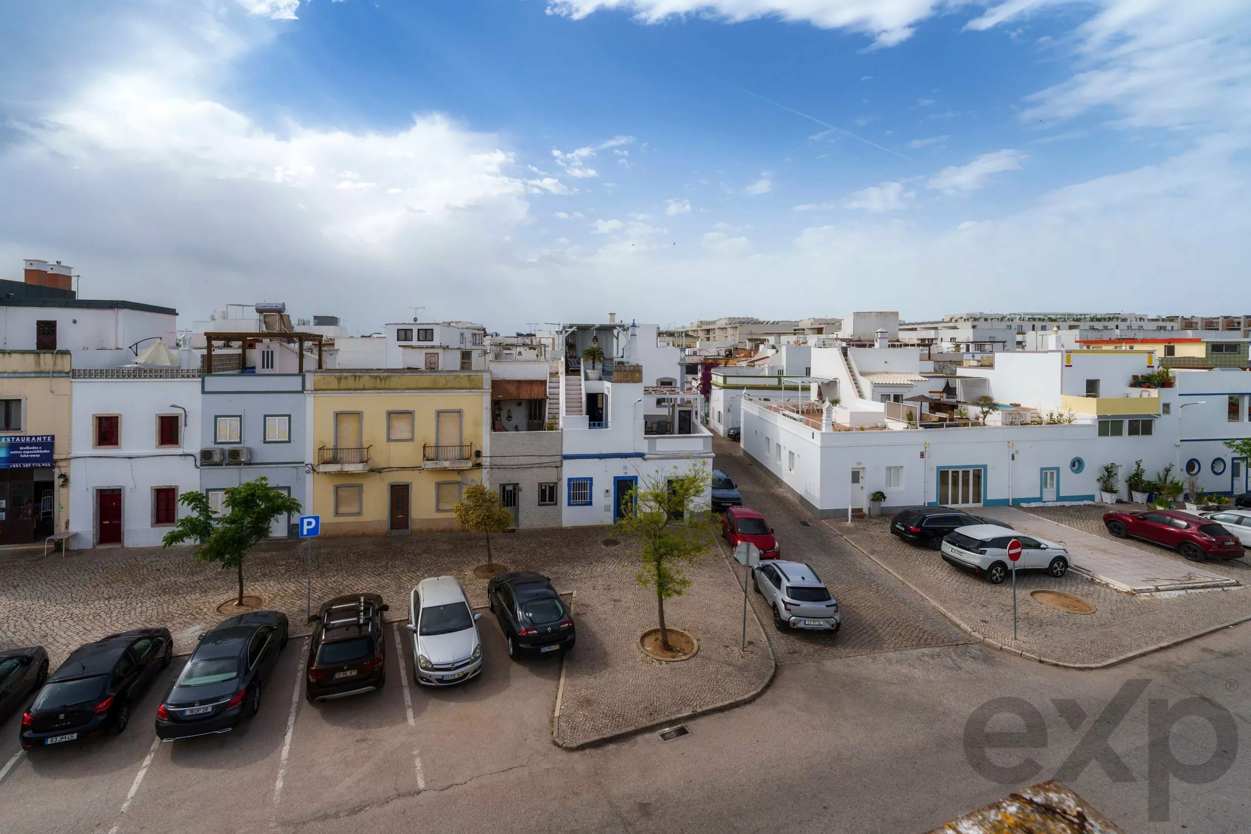 Wide view of a sunny coastal town square with white and pastel buildings, cobblestone streets, and parked cars beneath a blue sky.