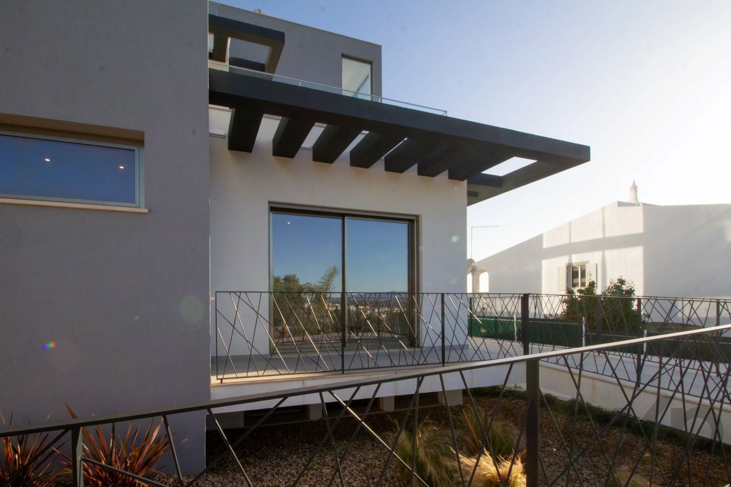 Modern two-story house exterior featuring a cantilevered black pergola, large glass windows, and a geometric metal railing.