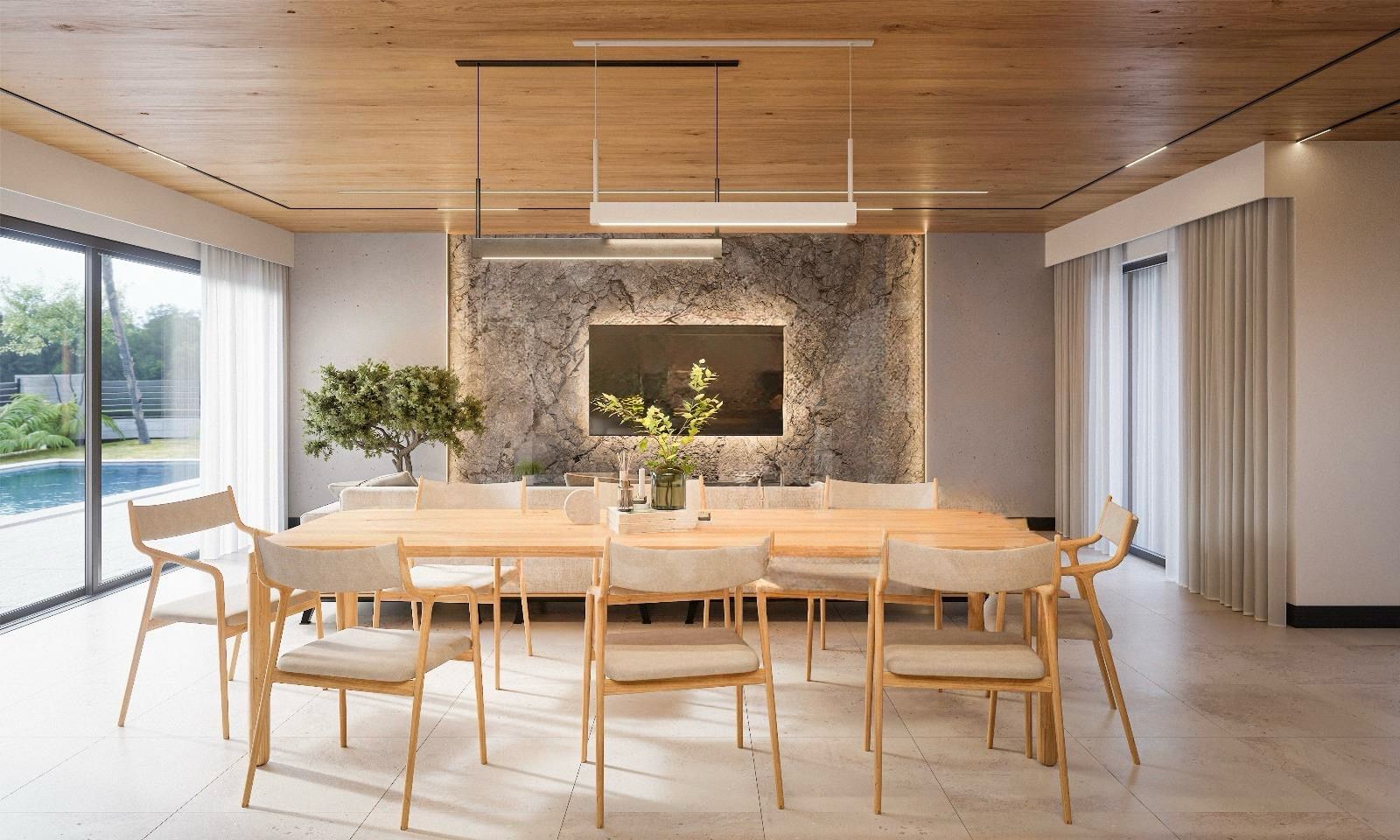 Modern dining room with a long wooden table and cream chairs, a stone wall with a mounted TV, and glass doors opening to a pool area outside.