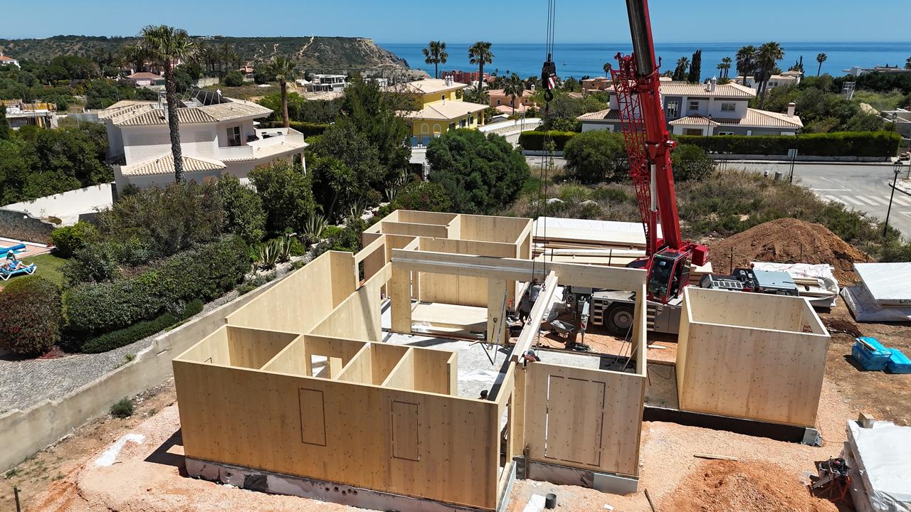 Wood-framed house under construction beside a coastal neighborhood, with a red crane lifting materials.