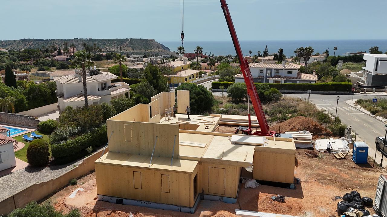 Wood-framed house under construction on a dirt lot; a red crane lifts materials with a coastal neighborhood and sea in the distance.