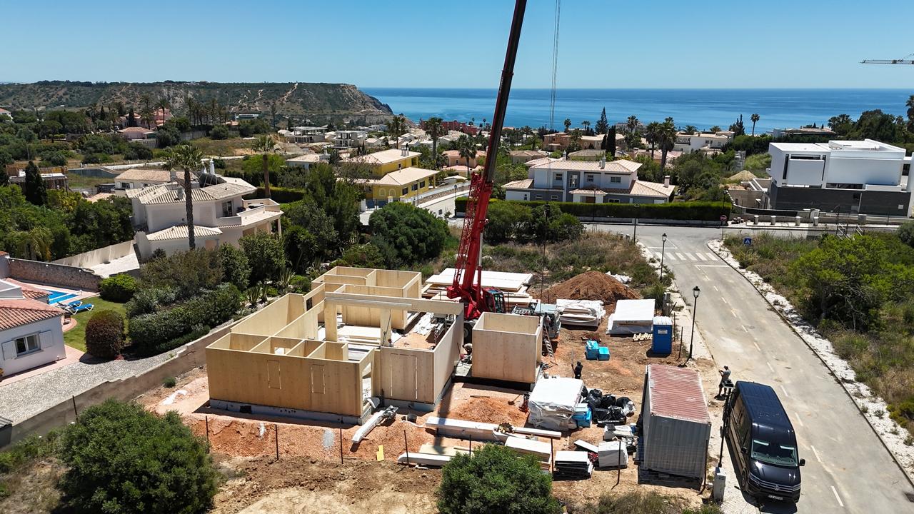 Aerial view of a construction site with wooden foundation walls and a red crane in a coastal suburban neighborhood near the sea.