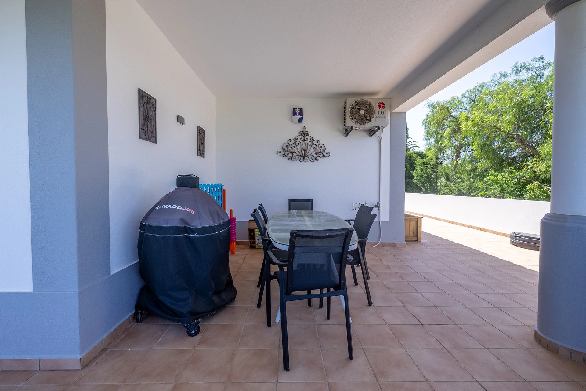 T5 Villa with pool in Ferrel 5mn from beach and Lagos centre – SunnySteve 18 Outdoor covered patio with a glass dining table and black chairs, grill under a protective cover, decorative wall art, and an LG outdoor AC unit; greenery beyond the railing