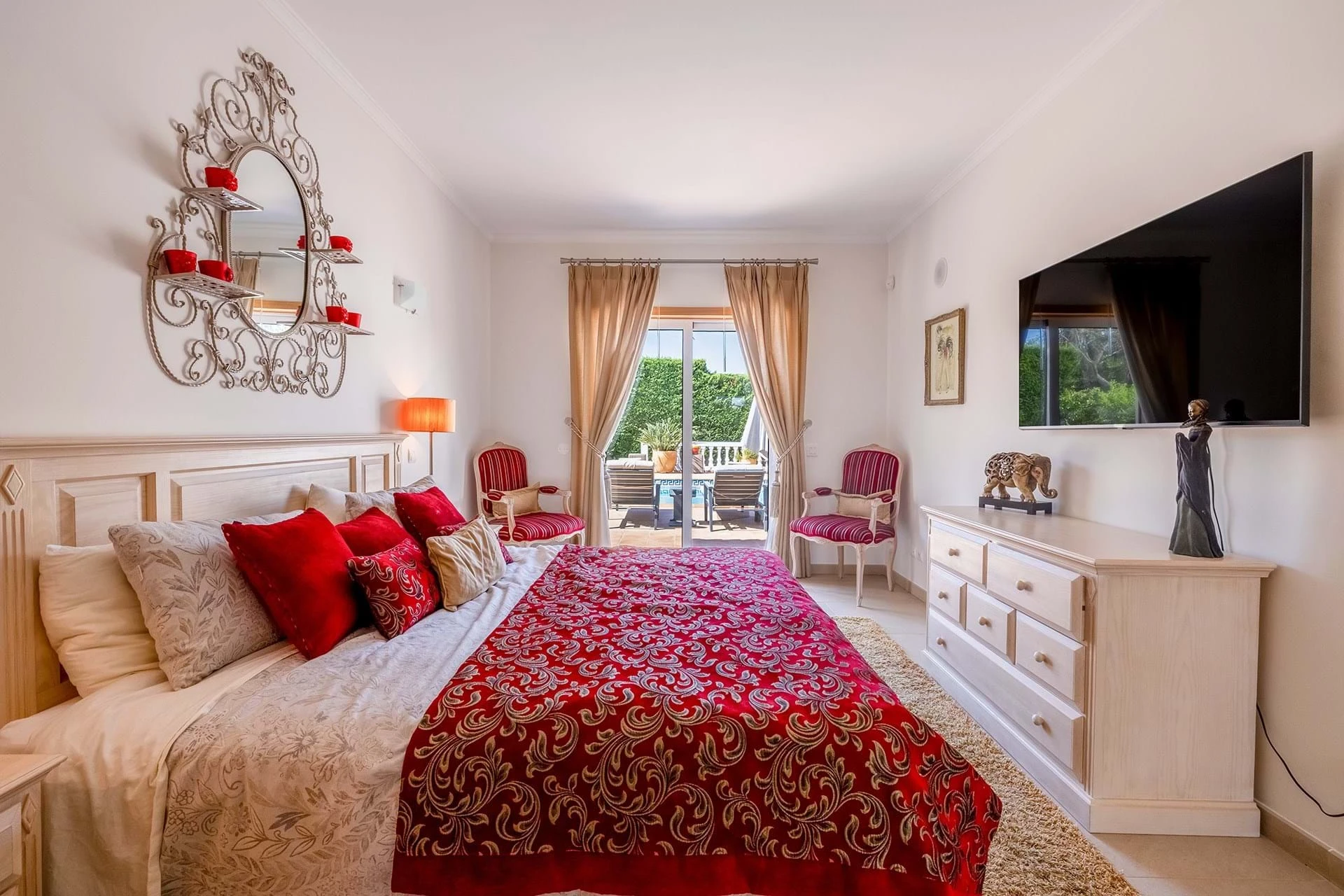 T5 Villa with pool in Ferrel 5mn from beach and Lagos centre – SunnySteve 20 Bedroom with a red patterned duvet and multiple decorative pillows, cream furniture, and an ornate metal wall mirror with red candle shelves above the headboard, near a balcony door with beige curtains.