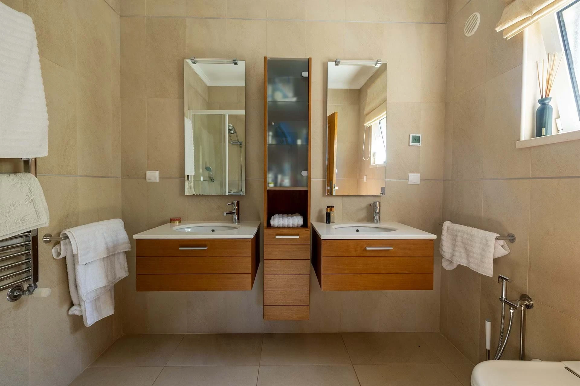 T5 Villa with pool in Ferrel 5mn from beach and Lagos centre – SunnySteve 24 Beige-tiled bathroom with two wooden vanities, white sinks, central glass-front cabinet, and large mirrors.